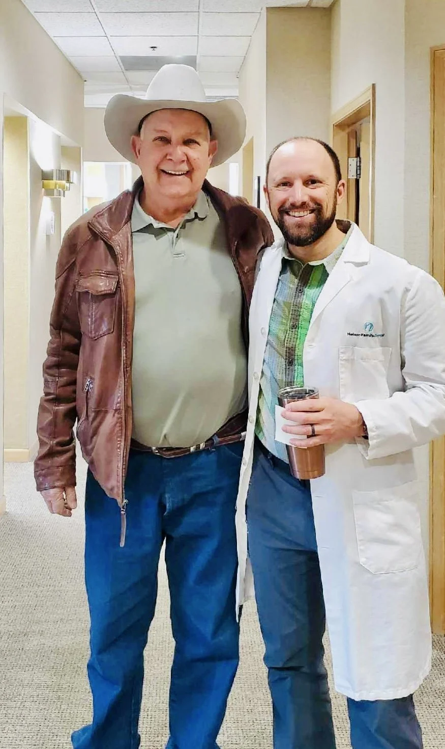 Two men smiling and posing together in an indoor hallway, one wearing a cowboy hat and leather jacket, the other in a white coat with a disposable cup in hand.  Dr. Chris Nelson with a friendly patient at Nelson Family Dental