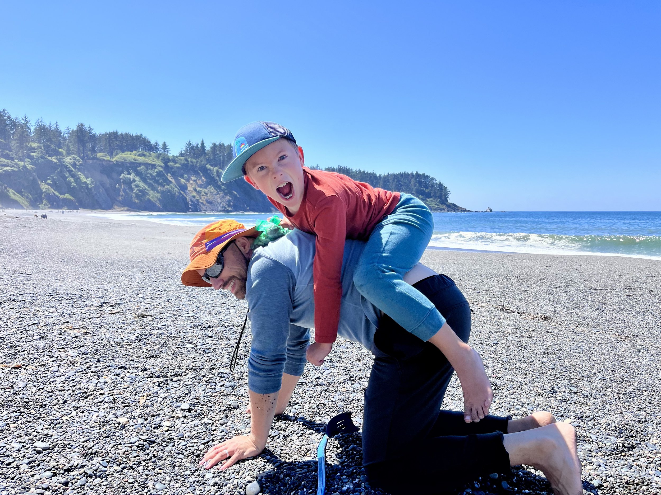 A young boy and an adult, both in athletic poses on a pebbled beach, with the boy on top in a mountain pose and the adult supporting him from below. The background features a blue sky, ocean, rocky cliffs, and trees.