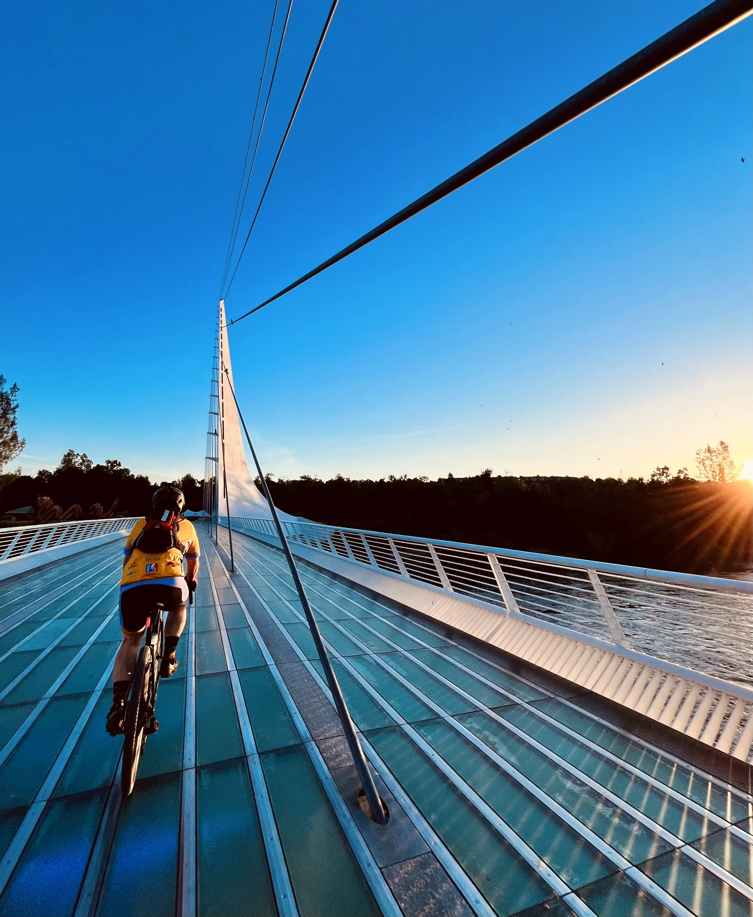 A person biking on a bridge with a modern, glass-covered surface, under cables supporting the structure, with a sunset sky in the background.