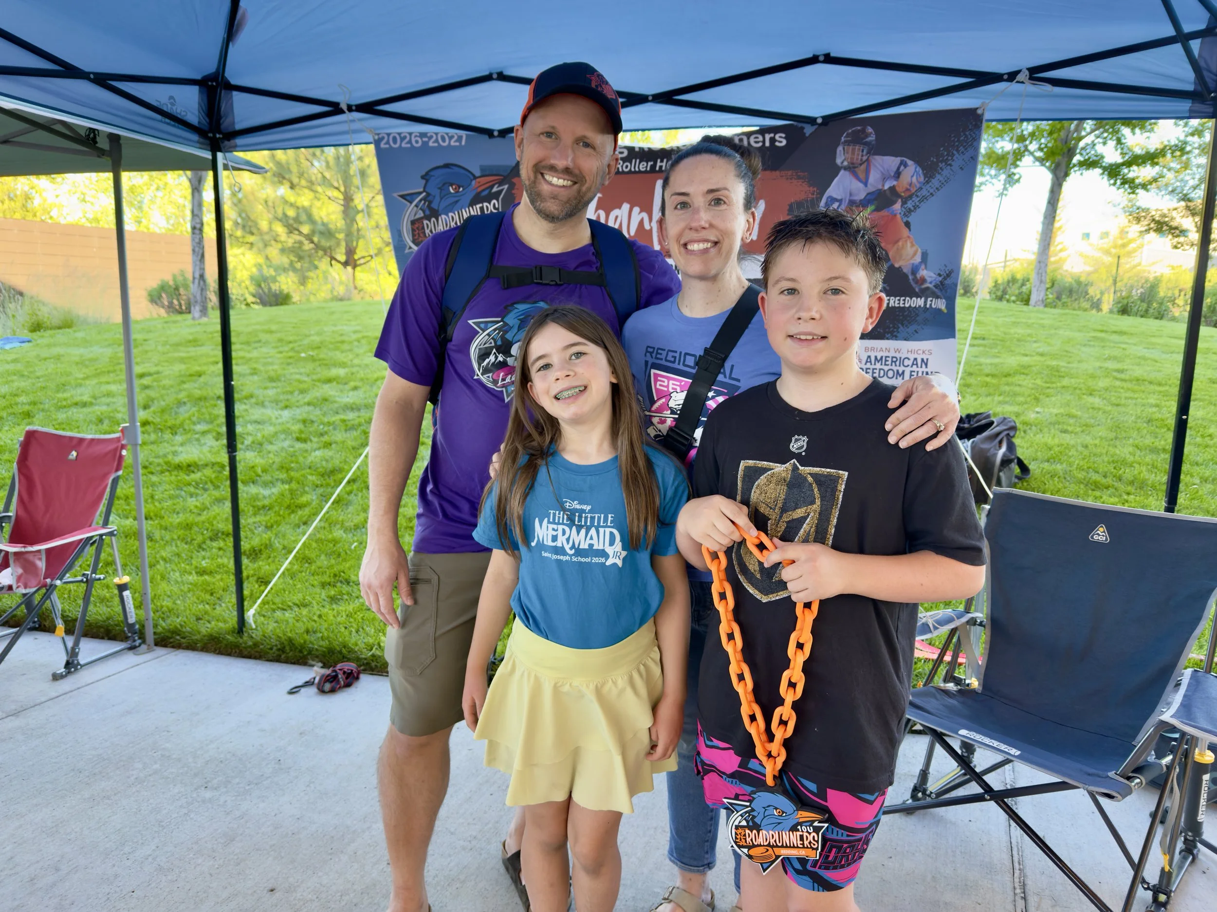 A group of four smiling people, two adults and two children, standing outdoors under a blue canopy tent during daytime. They are posing for a photo in front of a banner with a roller derby theme. The adults are wearing purple shirts, and the children