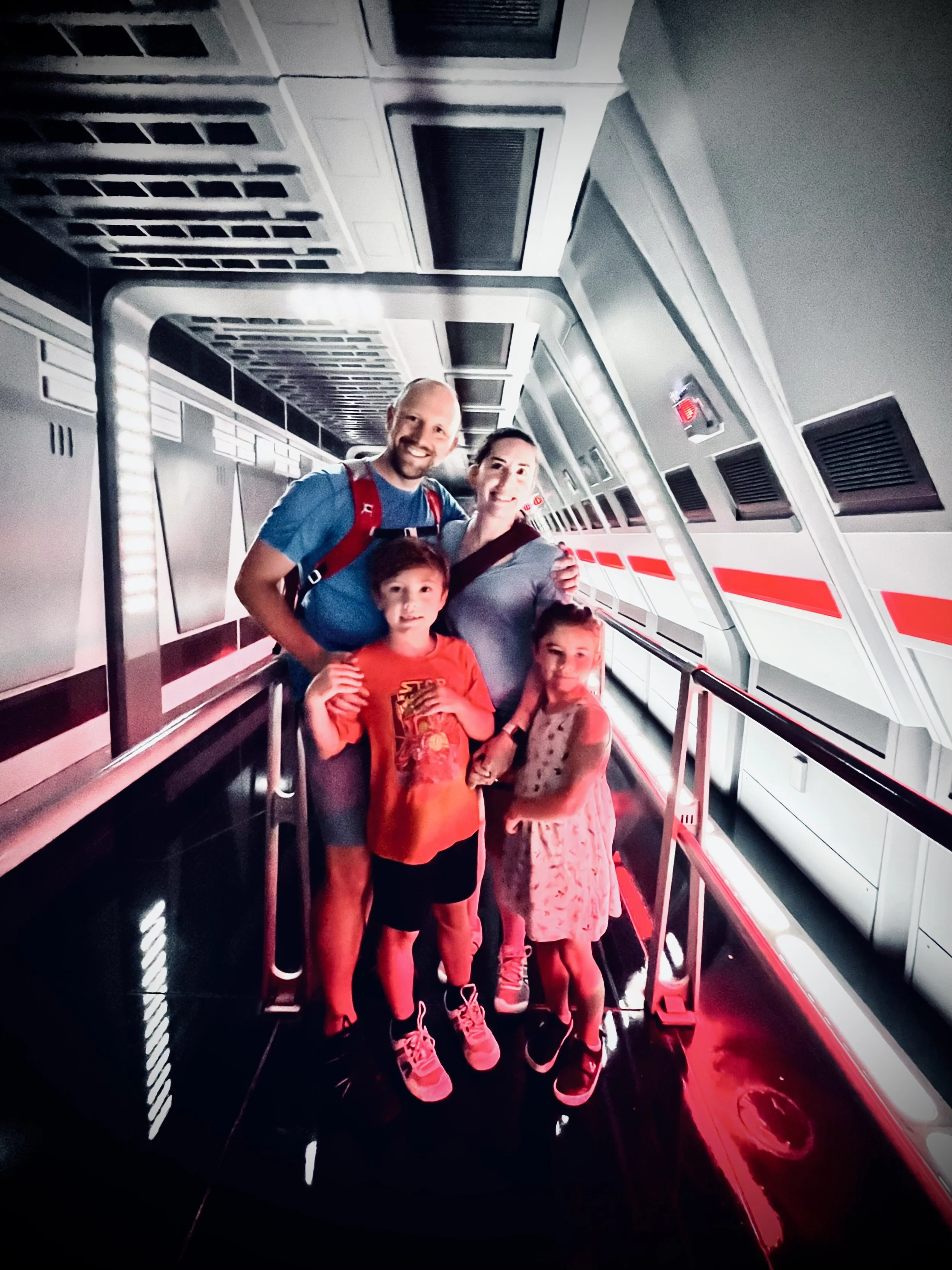 A group of five people, two adults and three children, standing inside a futuristic, metallic corridor with black and red lighting effects. The adults are smiling, with the man giving a thumbs-up and the woman standing close to the children, who are 