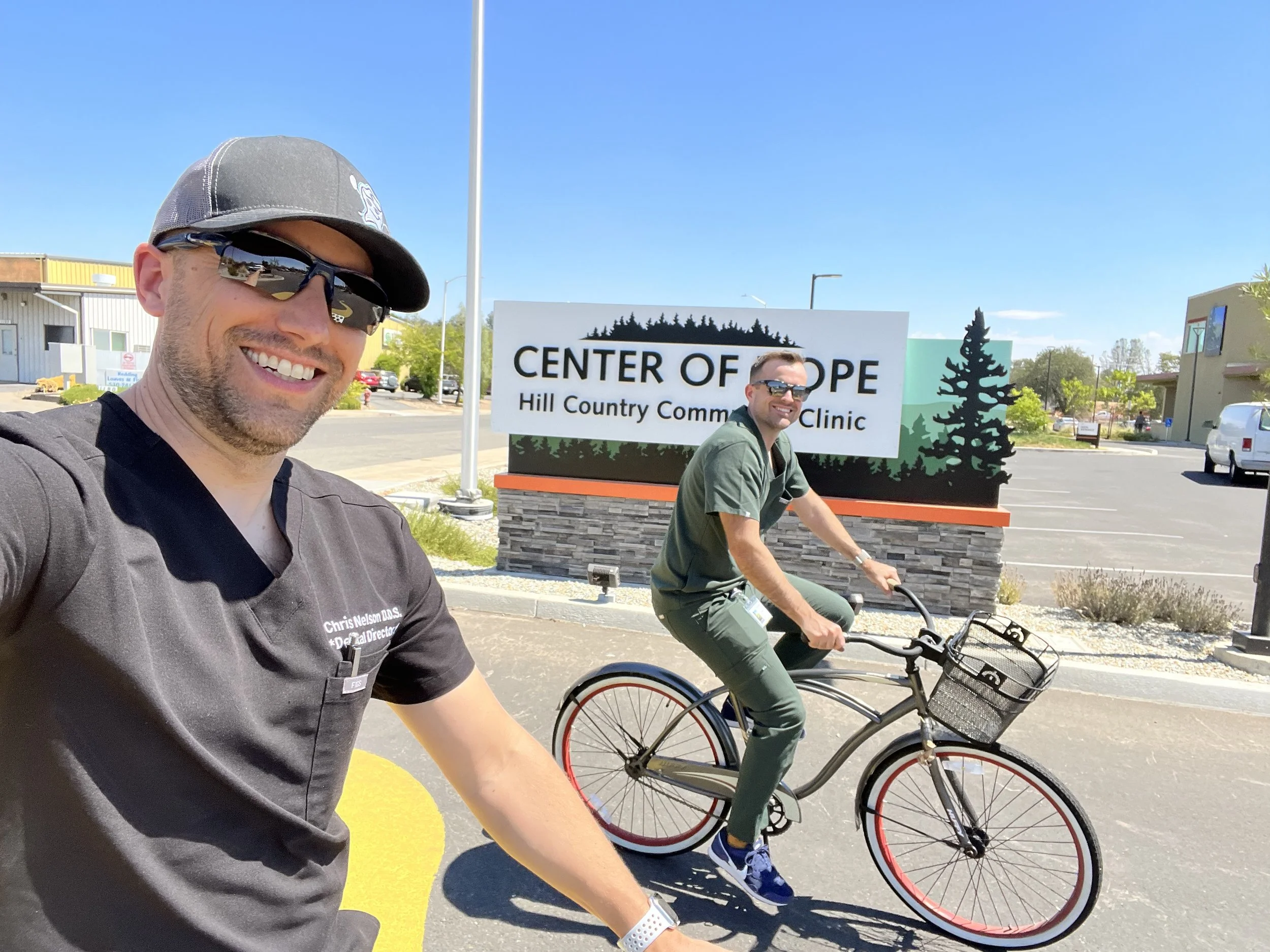 Two men riding bikes outdoors near a sign that reads 'Center of Hope, Hill Country Community Clinic' on a sunny day.