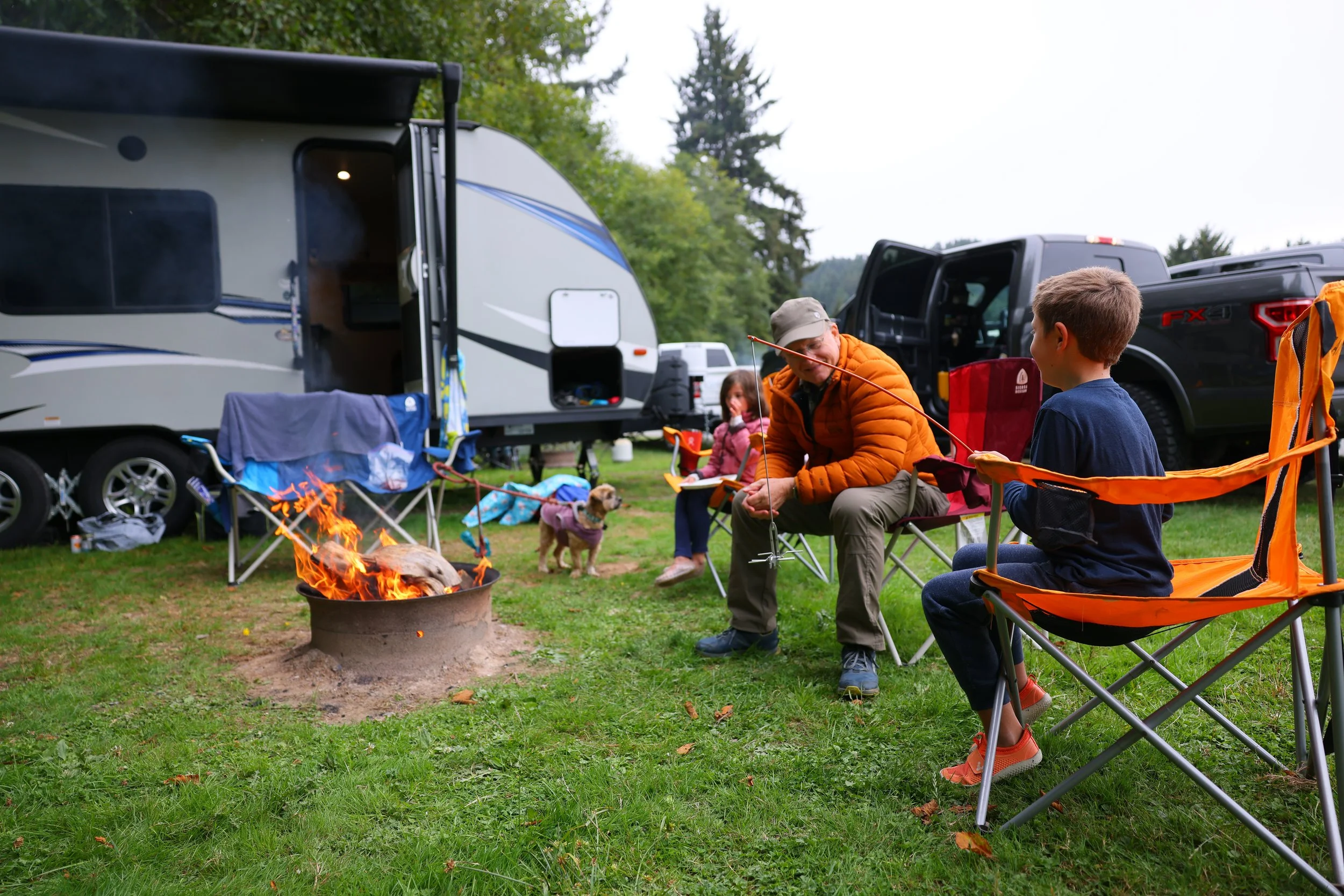 A family camping scene with a travel trailer, a fire pit with a fire, and several people sitting around, including children and an adult. The background features trees and parked vehicles.