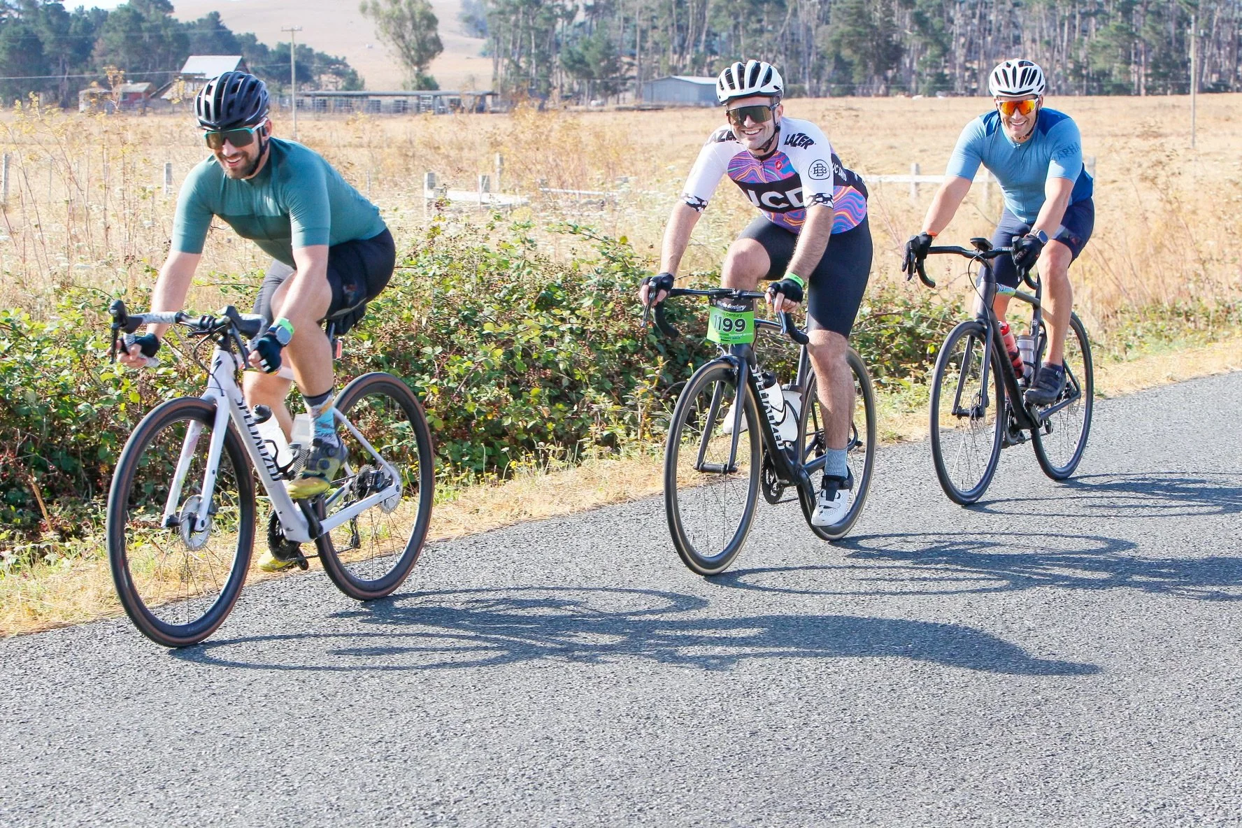 Three men riding bicycles on a rural paved road during daytime. They are wearing helmets and sunglasses, and are smiling. The background features fields, some trees, and distant structures.