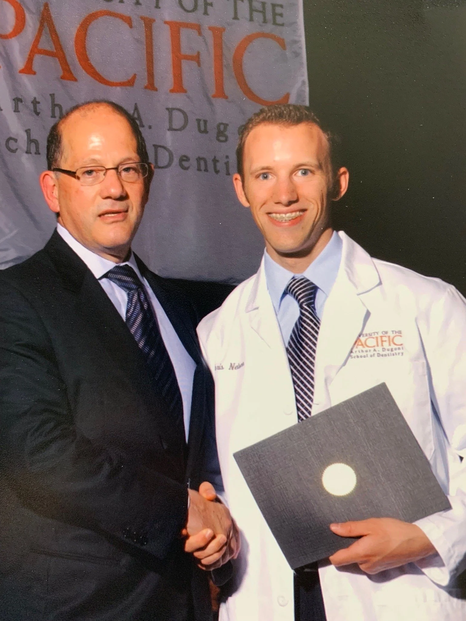 Two men, one in a suit and glasses, the other in a doctor's coat, shaking hands at a graduation or award ceremony with a banner behind them that reads 'University of the Pacific' and mentions 'Arthur A. Dugoni School of Dentistry'.