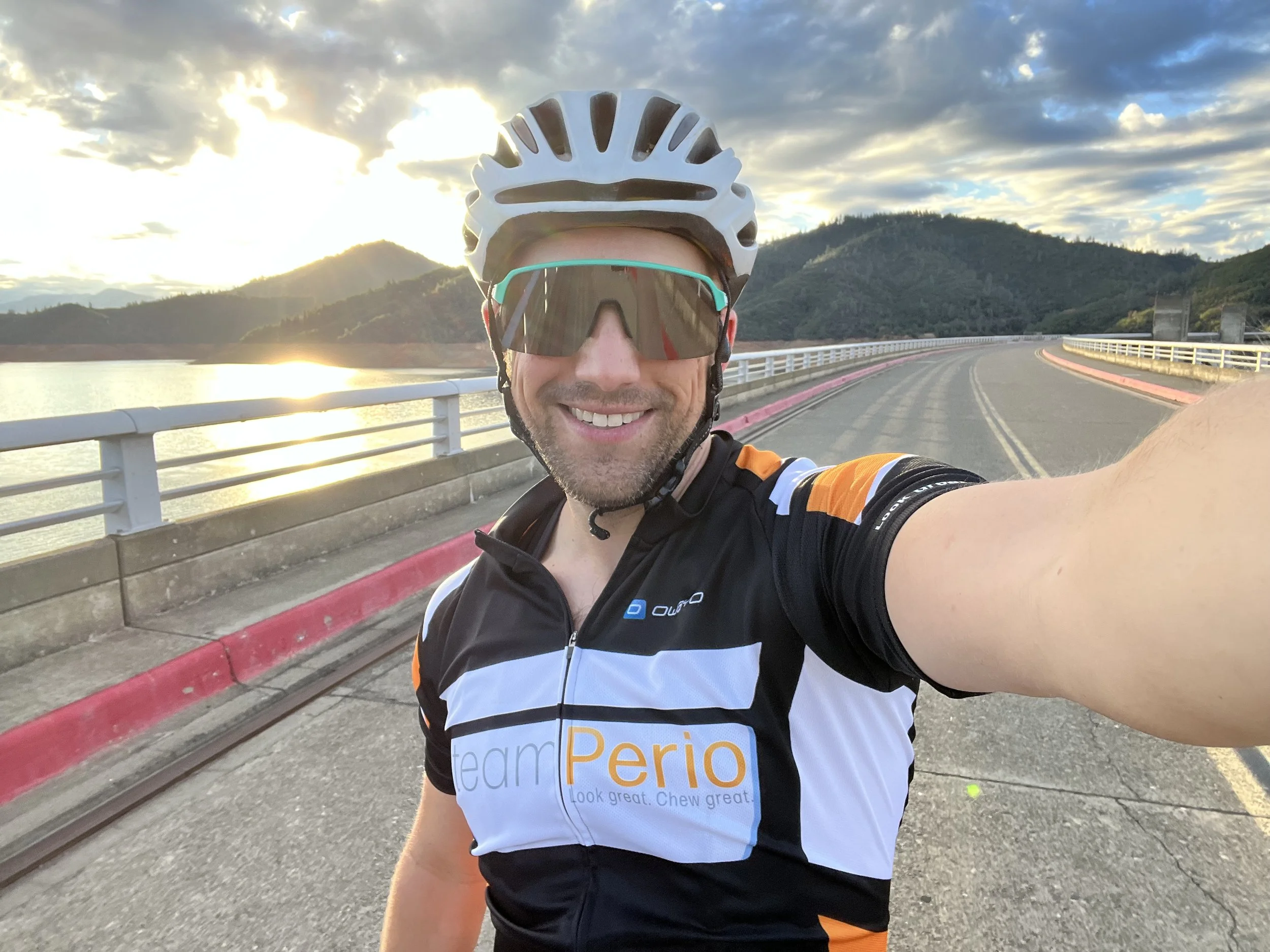 A smiling man wearing cycling gear, sunglasses, and a helmet, taking a selfie on a bridge with mountains and a partially cloudy sky in the background during sunset.  Team Perio Shasta Dam Redding Ca
