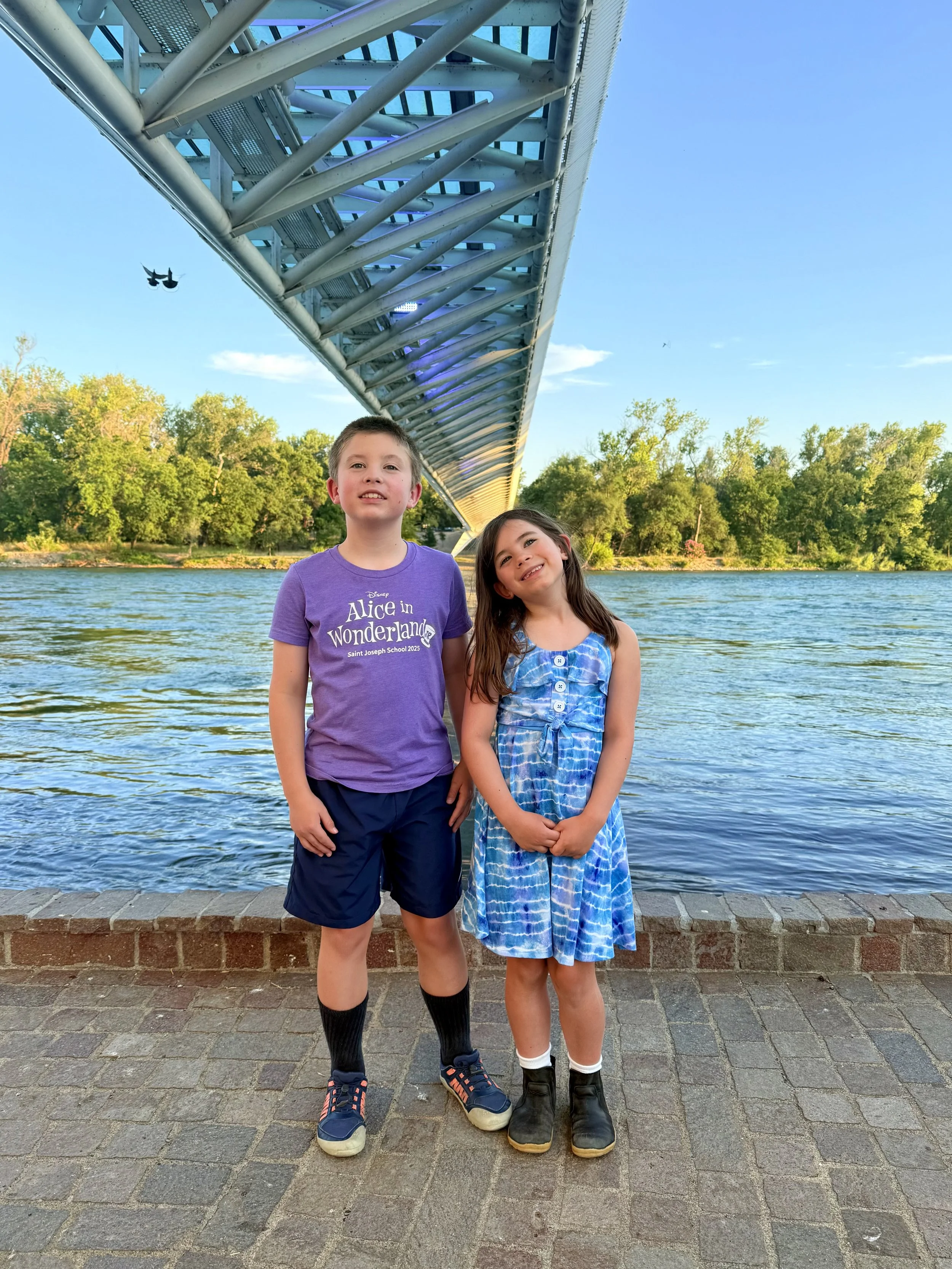 Two children, a boy and a girl, standing on a brick sidewalk near water, with a bridge overhead and green trees in the background. The boy is wearing a purple t-shirt, navy shorts, and sneakers, and the girl is wearing a blue dress with black boots. 