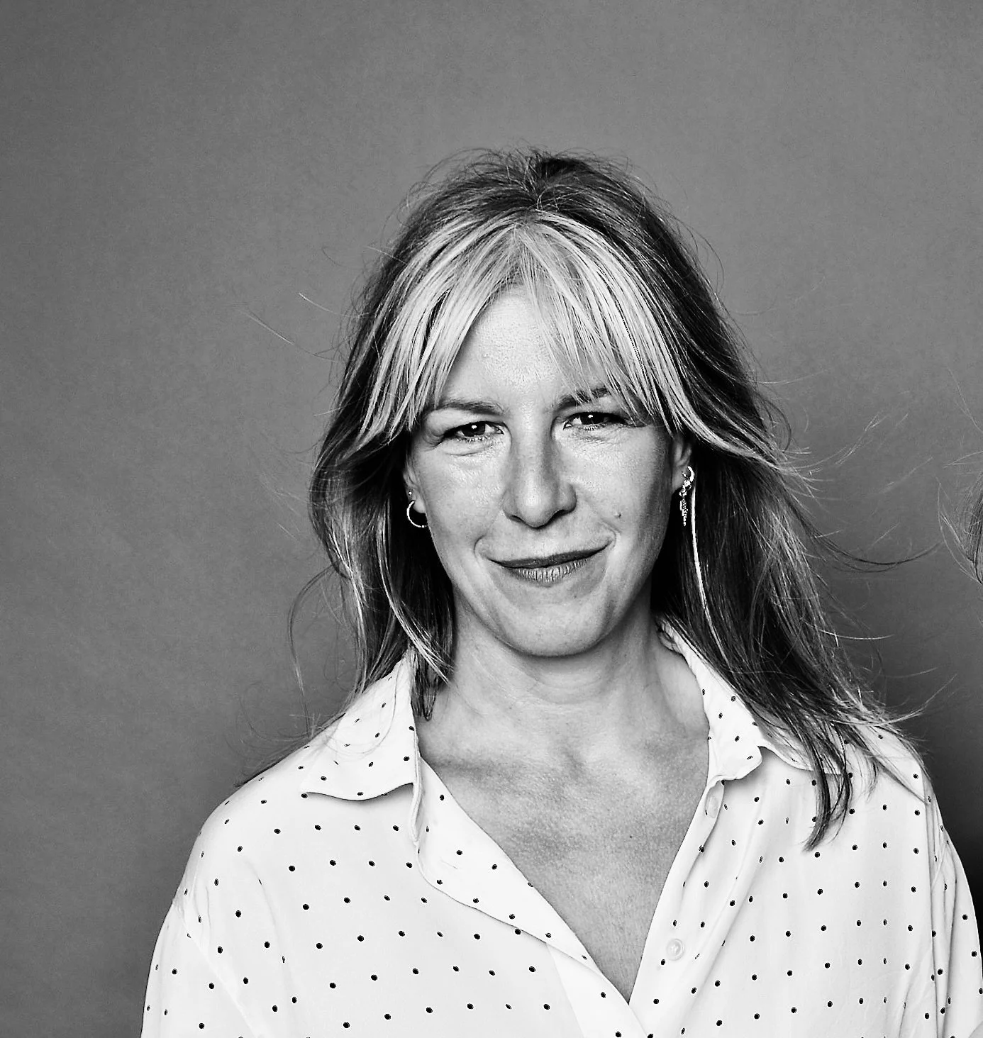 Black and white portrait of a woman with shoulder-length hair, wearing a dot-patterned shirt and hoop earrings, smiling slightly.
