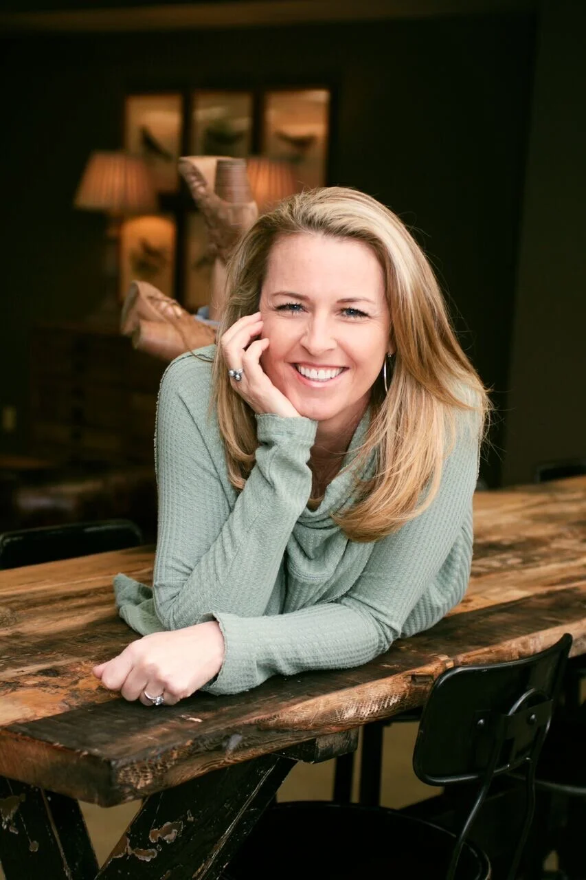 Image of owner Tracy Levinson on wood table wearing green shirt resting head on hand