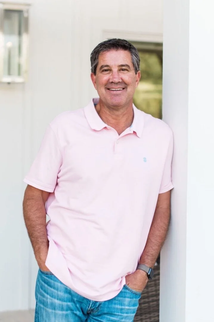 Owner Bruce Levinson leaning on white wall wearing pink shirt and blue jeans