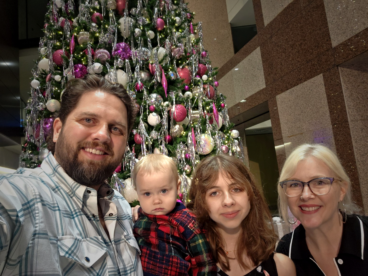 A family of four taking a selfie in front of a decorated Christmas tree. The man has a beard and is wearing a plaid shirt. The woman has blonde hair, glasses, and earrings. The young girl has curly red hair, and the young boy has short blonde hair and is wearing a plaid shirt. The background features a large Christmas tree decorated with pink, white, and silver ornaments, and string lights.