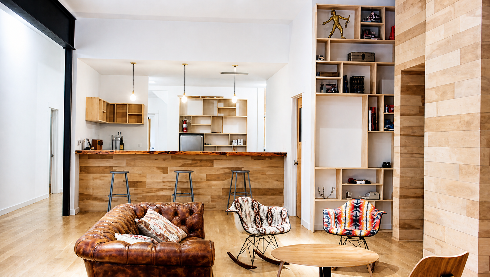 Interior of a modern living room with a wooden floor, a leather tufted sofa, patterned armchairs, a round wooden coffee table, and a built-in wooden shelving unit.