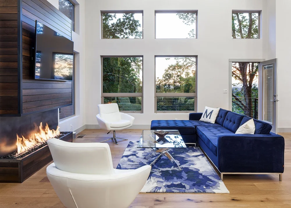 Double-height living room with a linear gas fireplace in a dark wood wall, a navy velvet sectional and tall windows onto oak trees