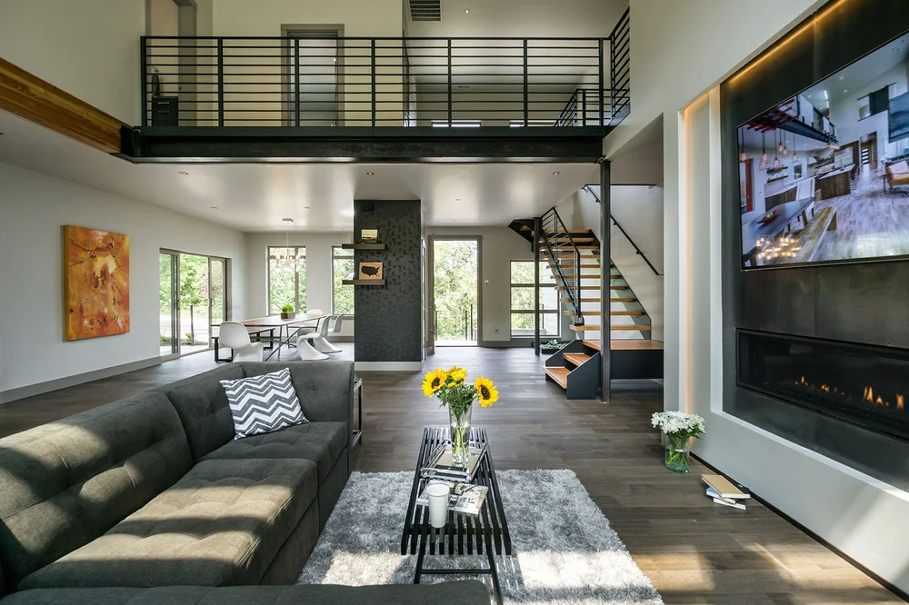 Double-height living room under a steel catwalk, with a floating stair and a linear fireplace