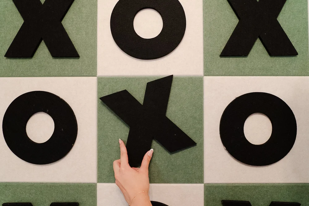 A hand placing a black X on a tic-tac-toe wall of green and white felt tiles