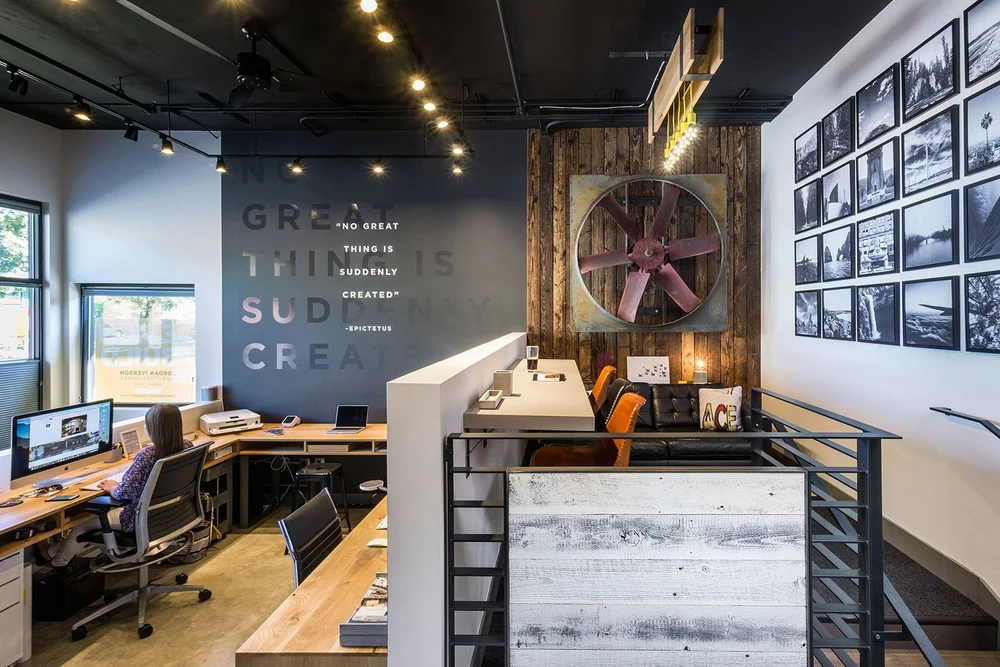 Studio office with oak desks, a charcoal quote wall, a vintage fan on reclaimed wood and a steel stair