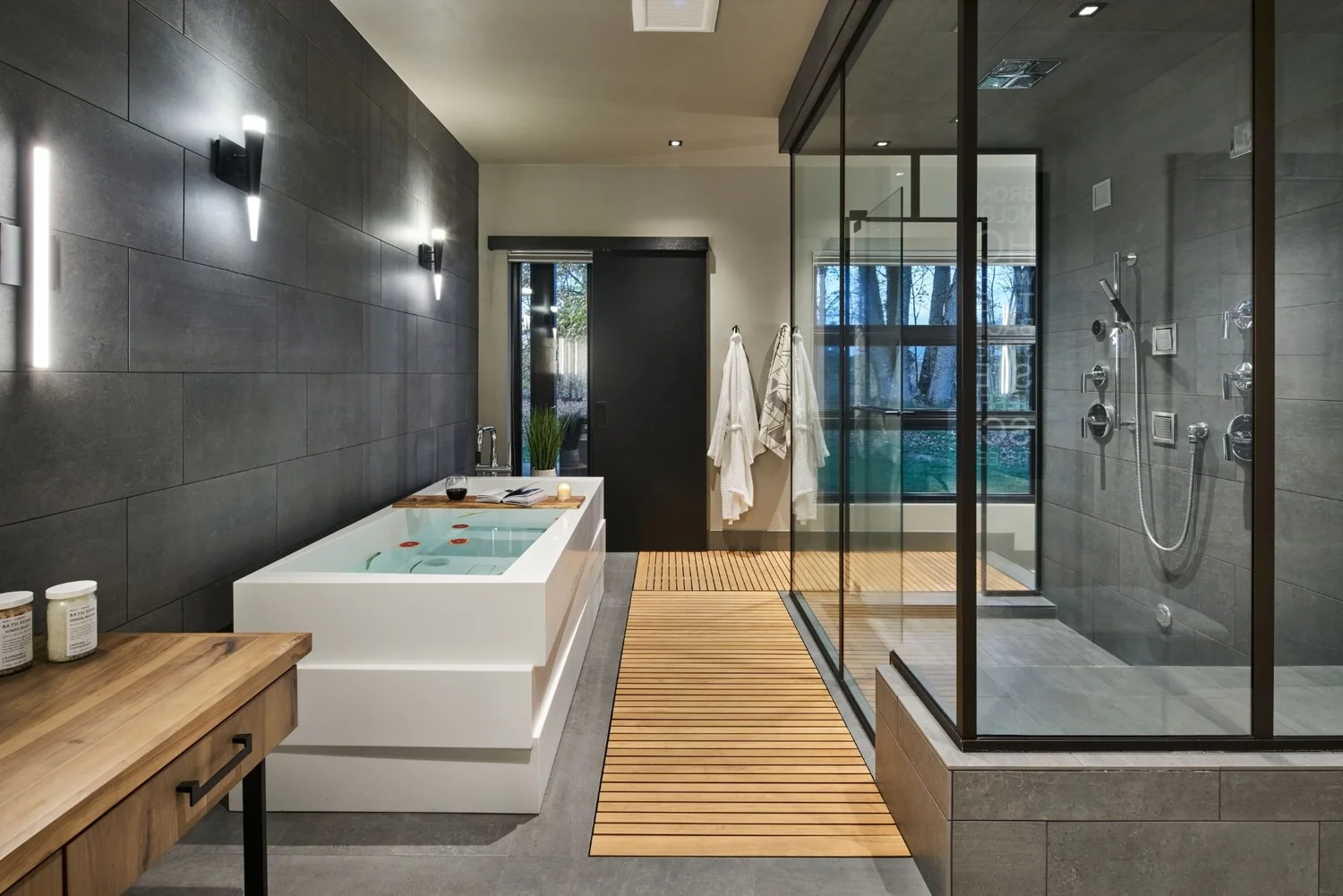 Primary bath with a white stepped soaking tub, dark tile wall, teak floor runner and a glass steam shower