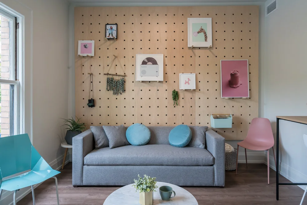 Gray sofa in front of a pegboard wall hung with art, with teal and pink chairs