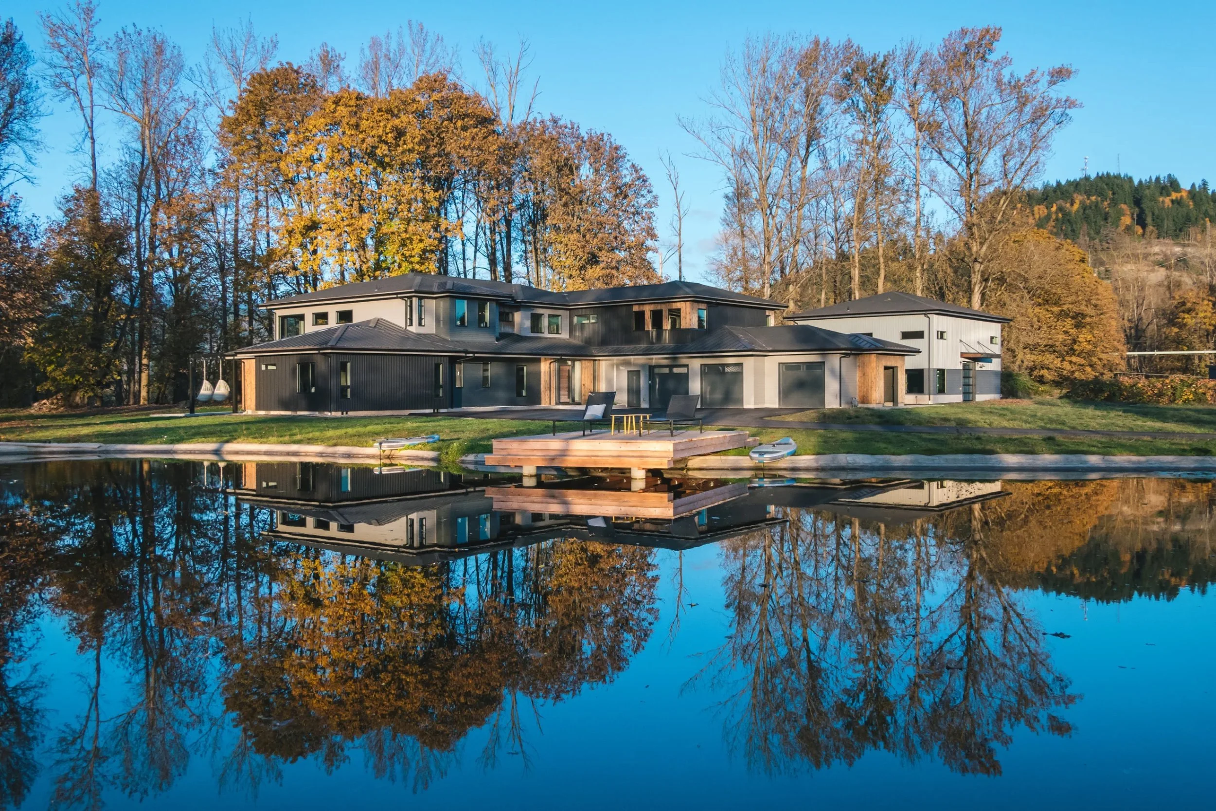 A dark-sided custom home with cedar accents and a floating dock, reflected in a still pond among autumn trees