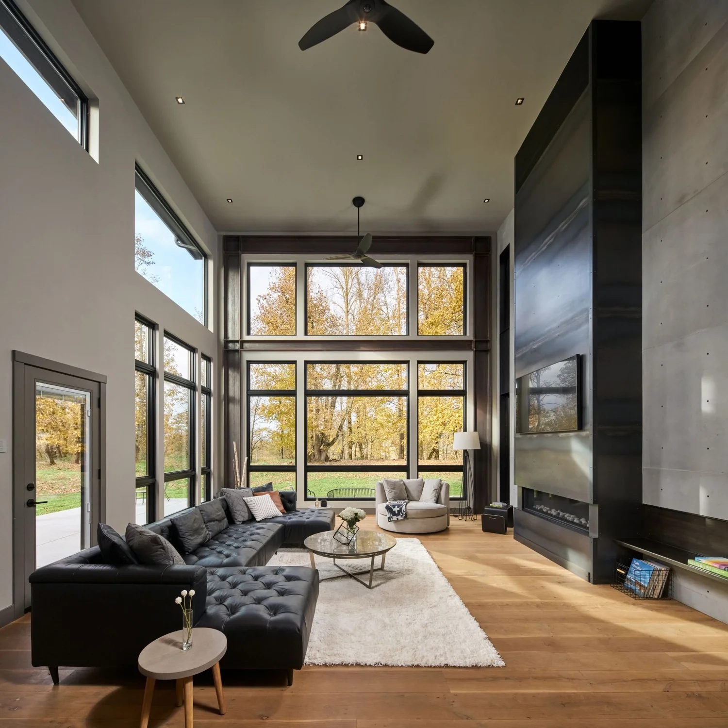 Double-height great room with a blackened steel fireplace wall, black leather sectional and tall windows onto autumn trees