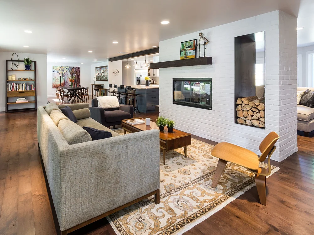 Open living room around a white brick double-sided fireplace with a firewood niche, looking through to the kitchen and dining room