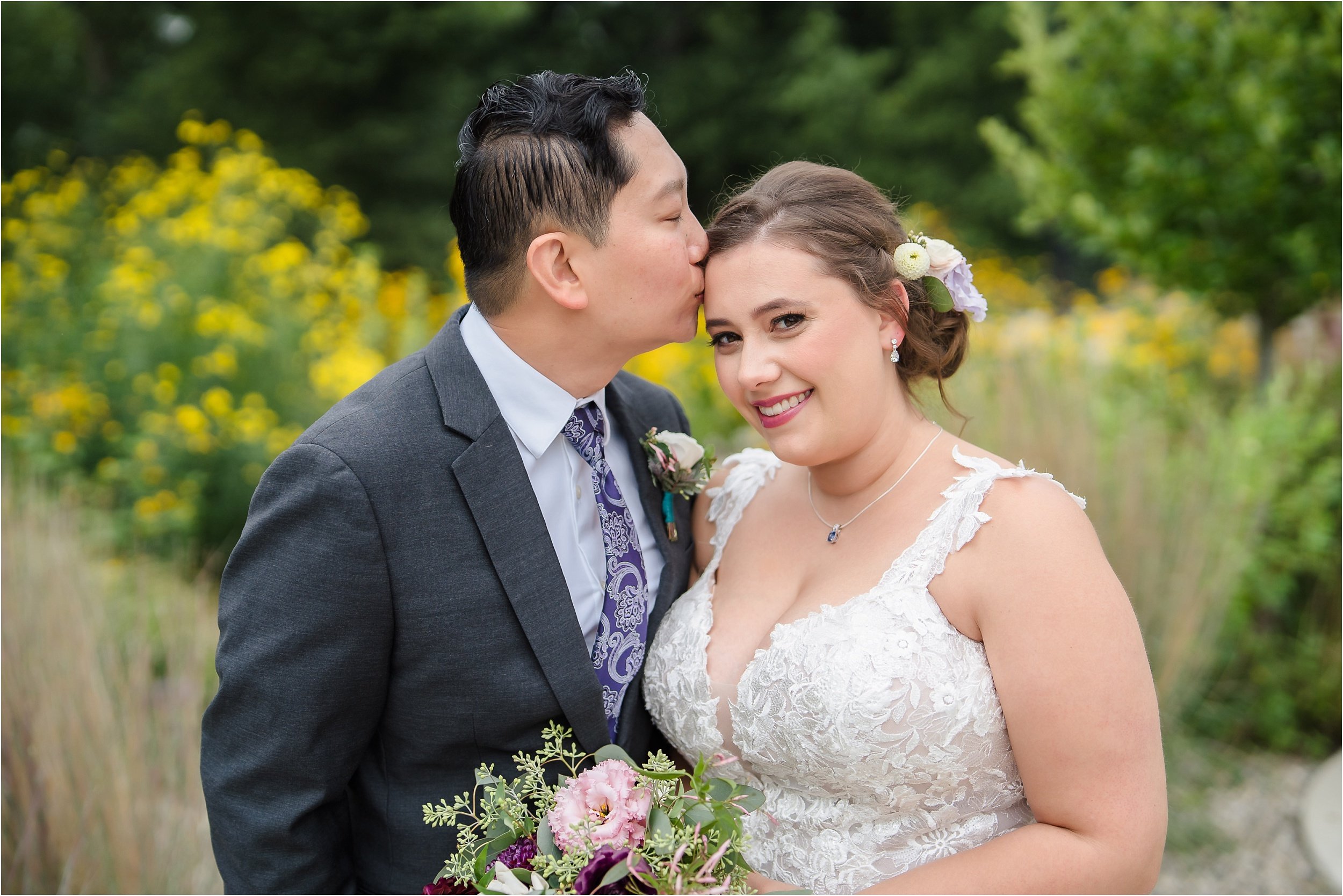  A groom wearing a floral tie kisses his bride on the forehead.  