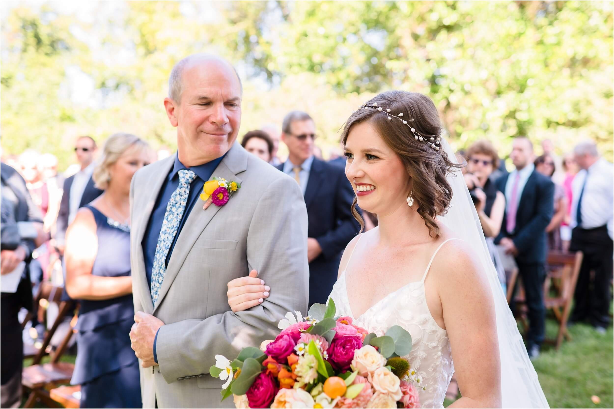  A dad looks lovingly at his daughter as they walk down the aisle towards her husband at their outdoor wedding ceremony.  