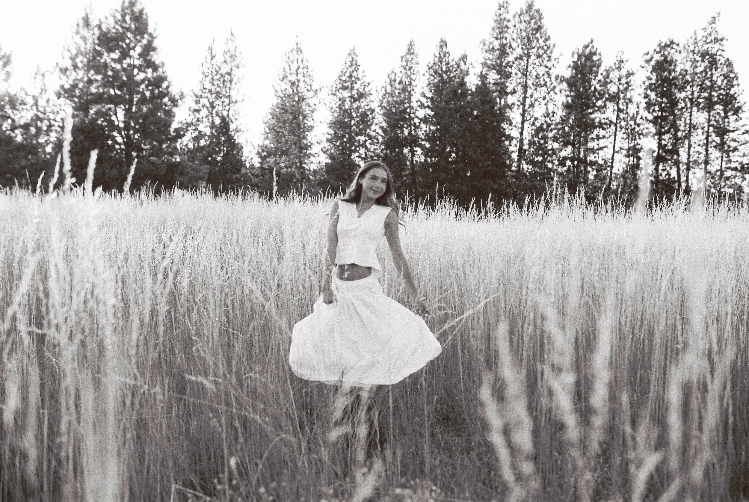 Spokane senior portrait black and white photo of a woman standing in a tall grass field, wearing a sleeveless top and a long skirt, with trees in the background.