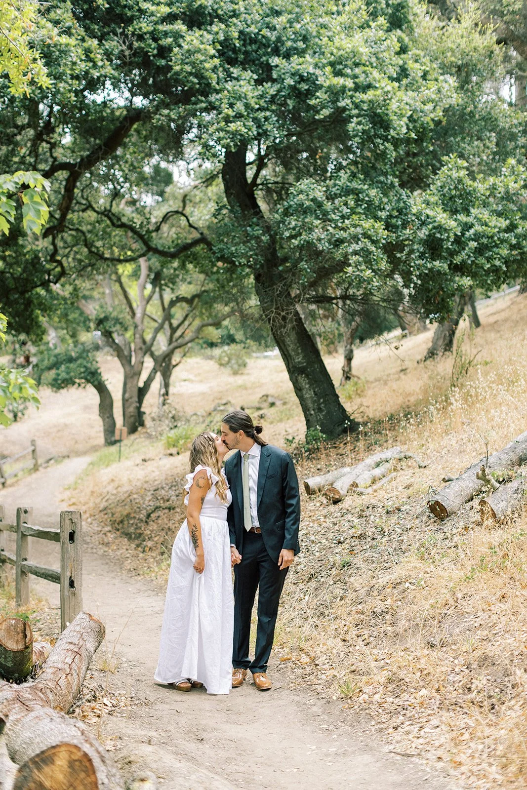 Couple walking hand in hand while kissing on a winding dirt trail through oak trees.