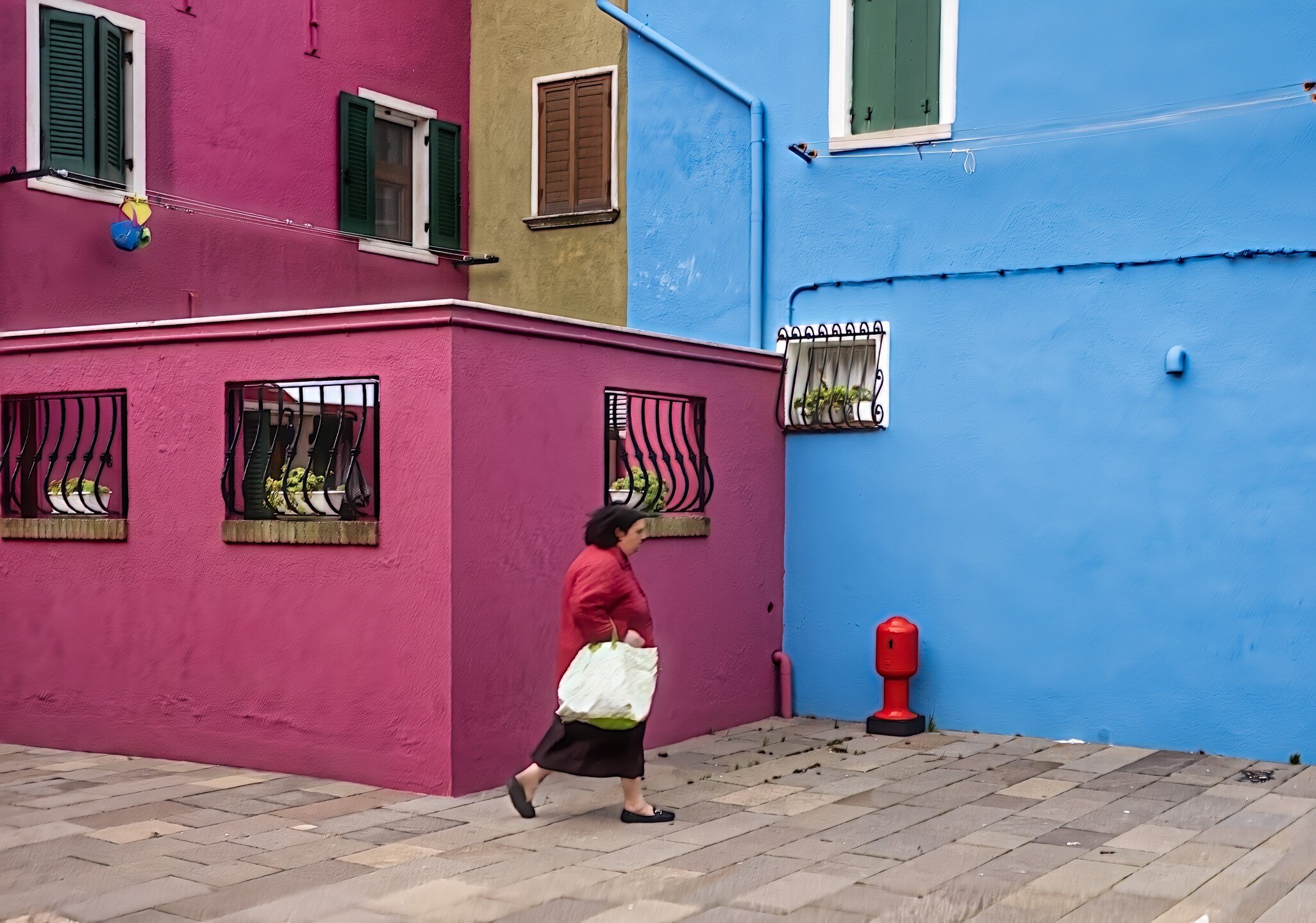 Traditionally captured through the lens of monochrome in my workshops, Burano's palette bursts to life in this rare departure from black and white. A local woman, clothed in a red that mirrors the vivid walls, carries the day's shopping, adding a pul