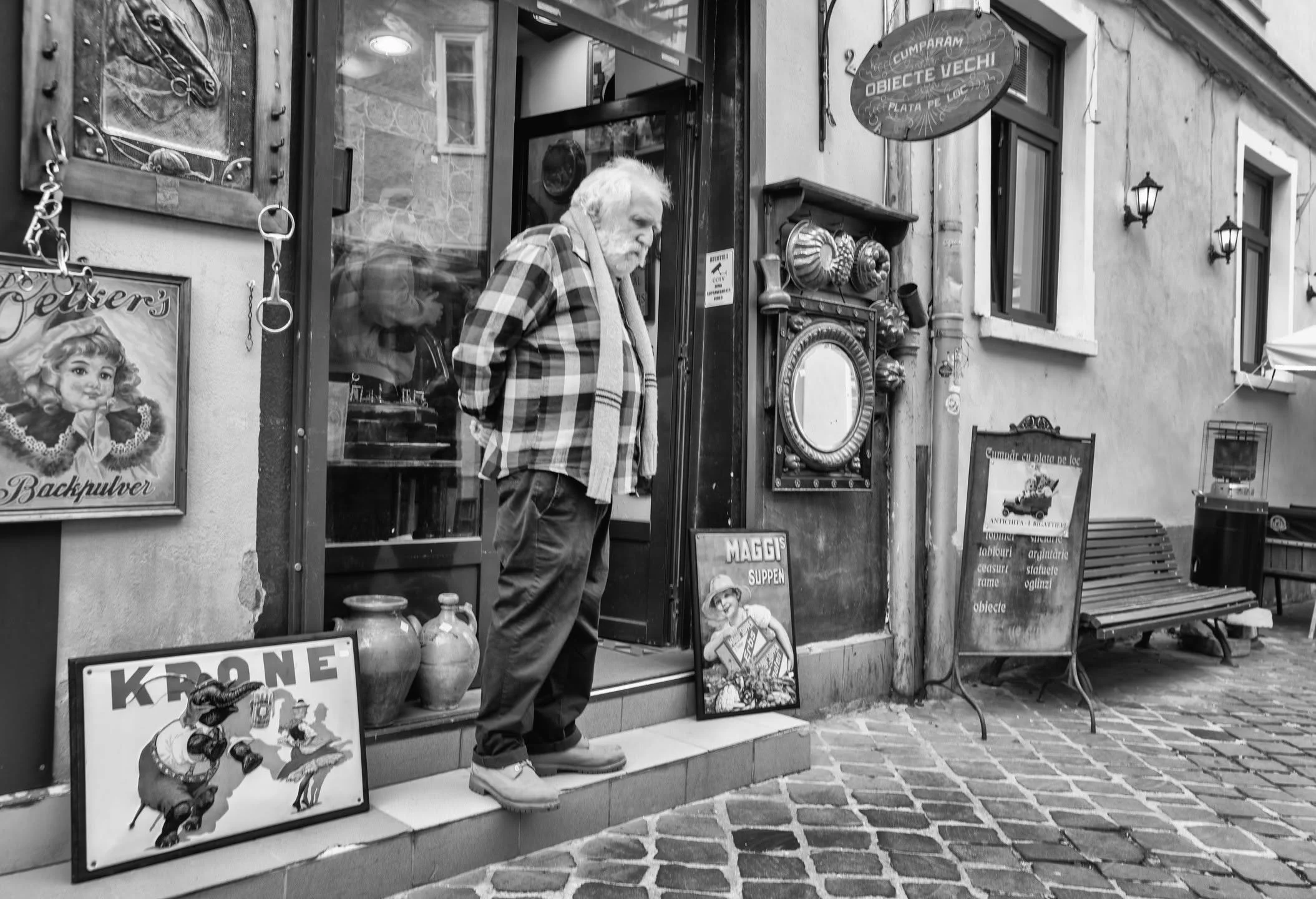 In Brasov there is always a moment like this, a quiet pause outside an antique shop where time feels heavier than the objects on display.
He steps out, hands behind his back, as if inspecting the street the same way he inspects the artefacts he lives