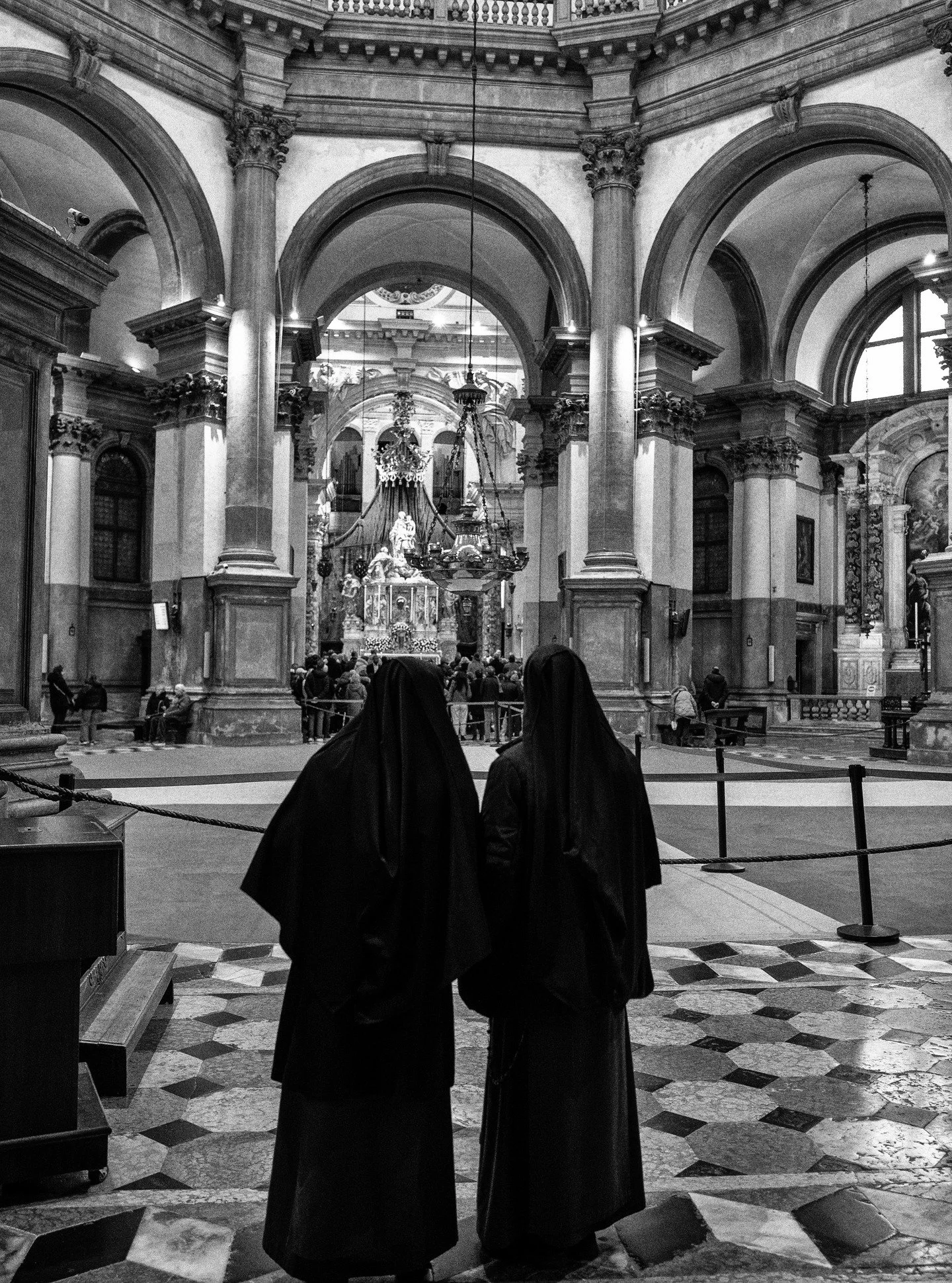Two nuns observe a service at the Basilica di Santa Maria della Salute in Venice, framed by the baroque arches and marble floor of the historic church, Italy.