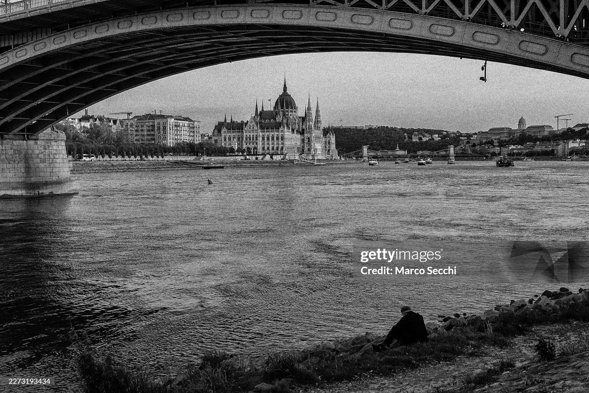 The Hungarian Parliament building is seen across the Danube River with a man sitting on the bank, in a monochrome composition. The Neo-Gothic landmark, completed in 1904, remains one of the most recognisable architectural symbols of Budapest and a fo