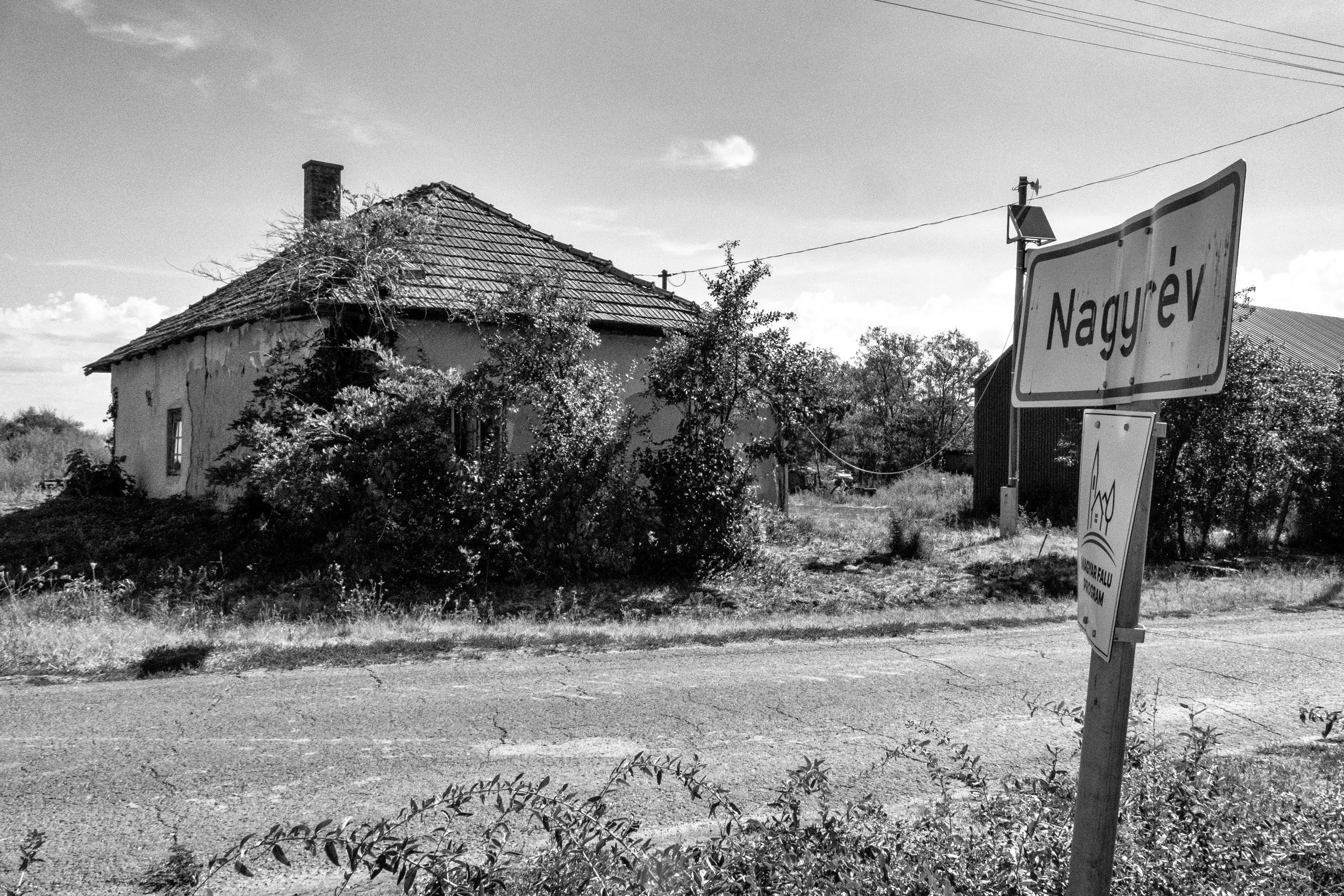 At the edge of Nagyr&eacute;v, the village announces itself quietly. A weathered house is slowly being reclaimed by the vegetation, while the empty road leads into a place that seems to hold more than it reveals.

I have become increasingly obsessed 