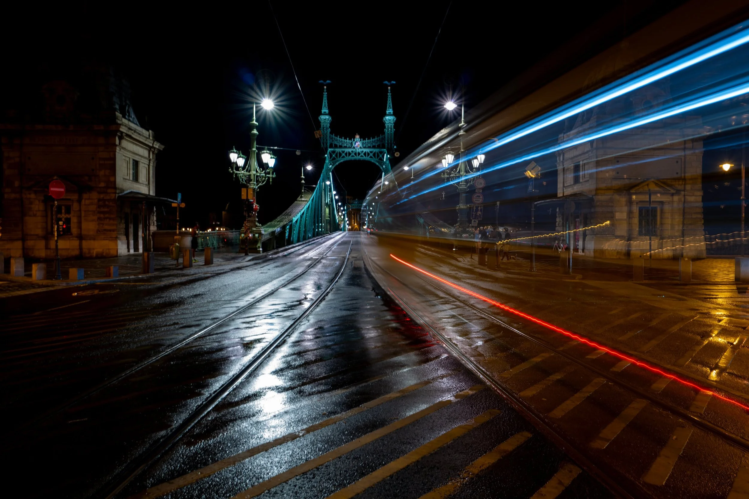 Budapest after rain, when the city seems to hold the light a little longer. 

A tram crosses Liberty Bridge, dissolving into blue and red streaks as the wet rails pull the eye toward Buda.

For a few seconds, the traffic, the river and the night fall
