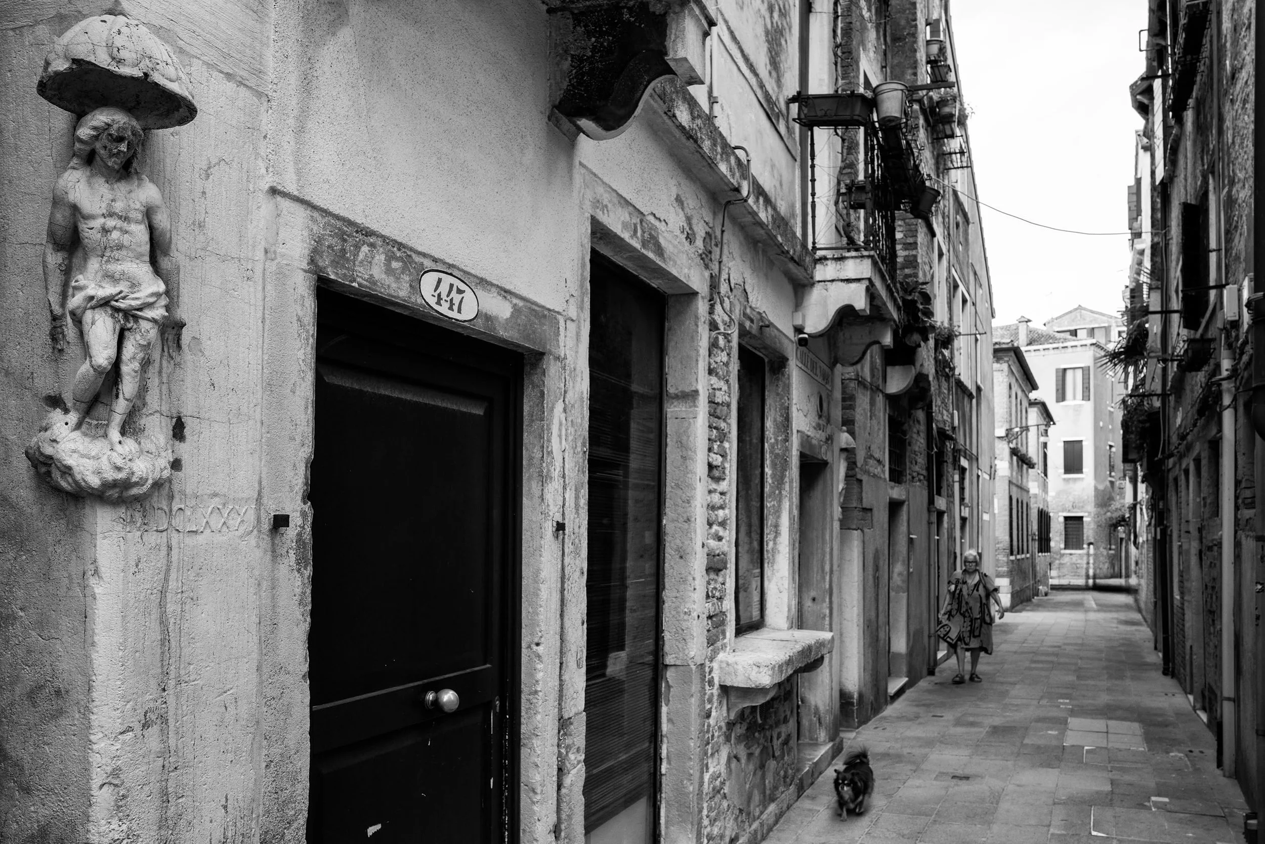 Venice keeps its magic in the space between the grand view and the ordinary day.

A man passes beneath the old lamps. A woman crosses a bridge above a blunt confession on the wall. A weathered saint watches over a narrow calle while a small dog trots