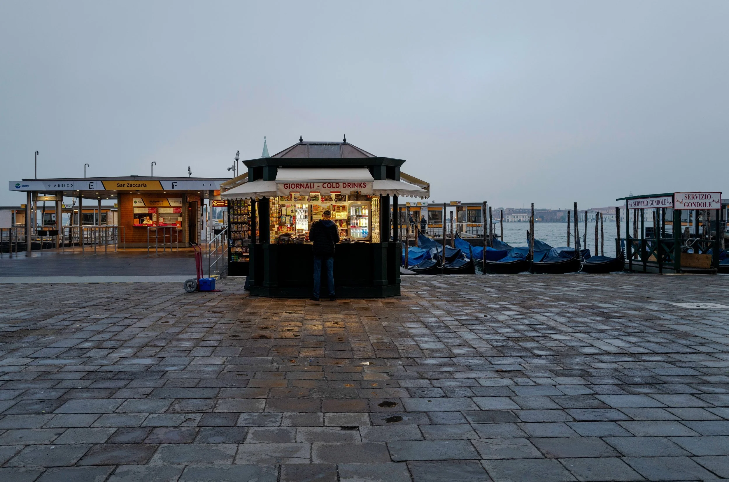 Early morning in Venice, and the brightest thing on the waterfront is a newspaper kiosk.

The gondolas are still covered. The lagoon and sky are almost the same blue-grey. One person stands at the counter, framed by the warm light spilling onto the d