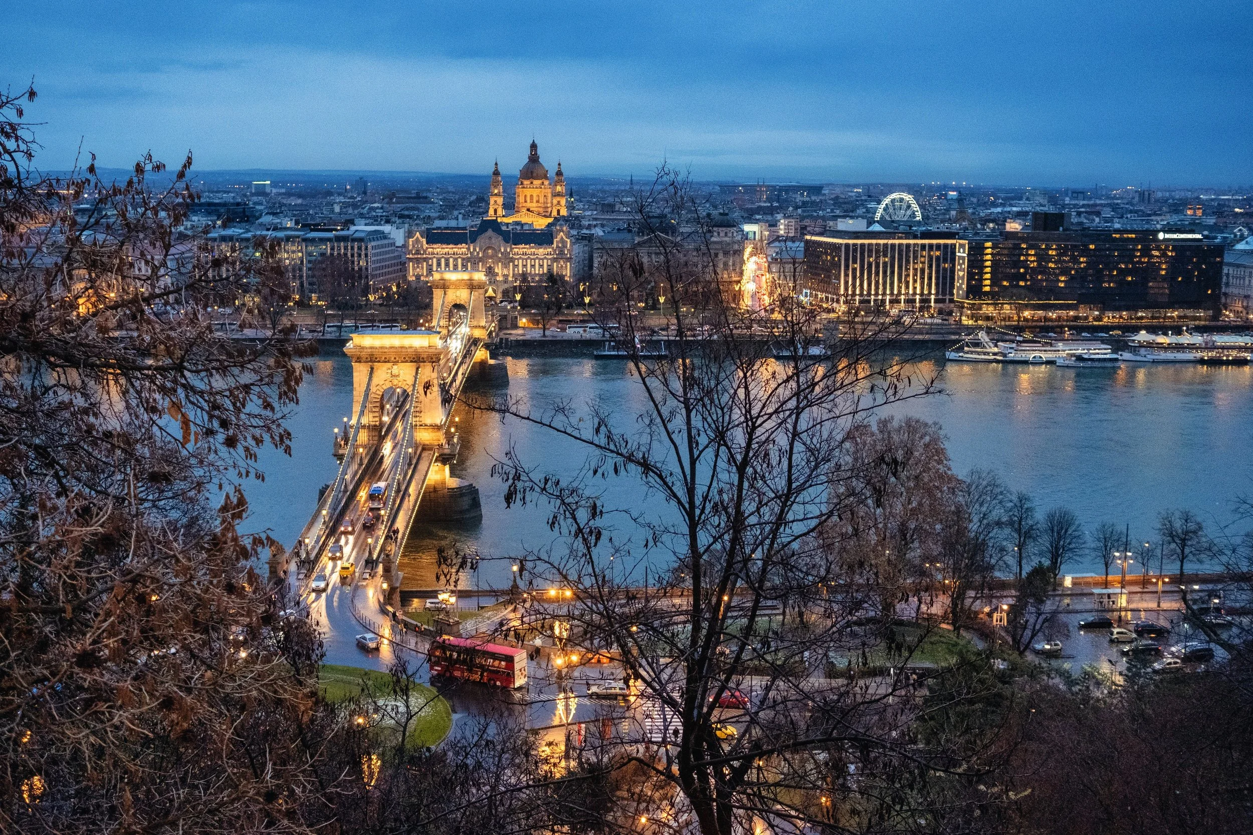 Budapest at dusk has a lovely balance, when the sky still holds its blue and the city lights begin to warm the river. Through the bare branches, traffic crosses the bridge, and reflections gather on the wet streets below. Even in a familiar view, som