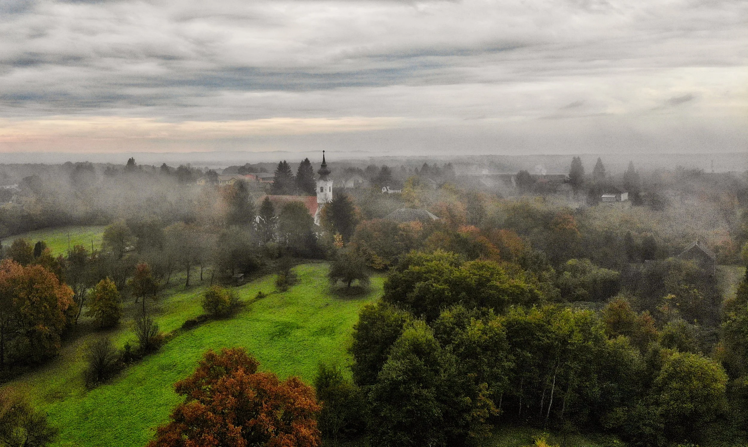 Autumn in Szentgy&ouml;rgyv&ouml;lgy, in the Őrs&eacute;g. Mist softens the edges of the village, but the green of the fields holds its own. Among the trees and scattered roofs, the church tower gives the eye somewhere to settle.

What draws me to th