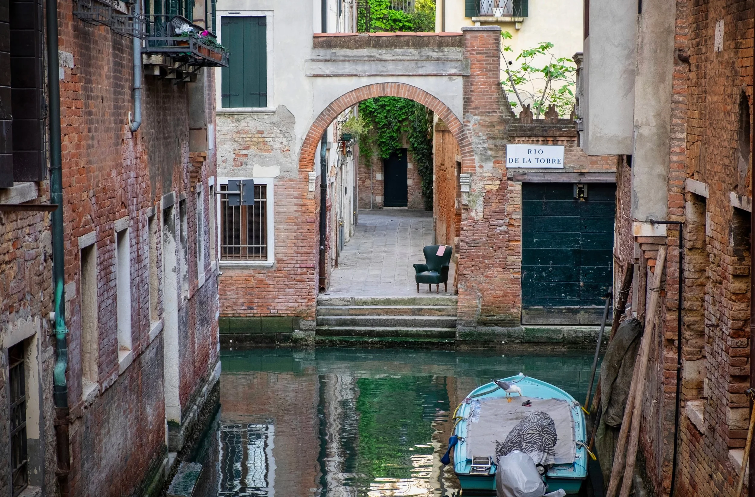The empty chair across the canal is the detail that holds my attention here. The brick archway gives the eye somewhere to go, but the chair gives it somewhere to stay. Around them, the worn walls, green water and reflections make a photograph from an