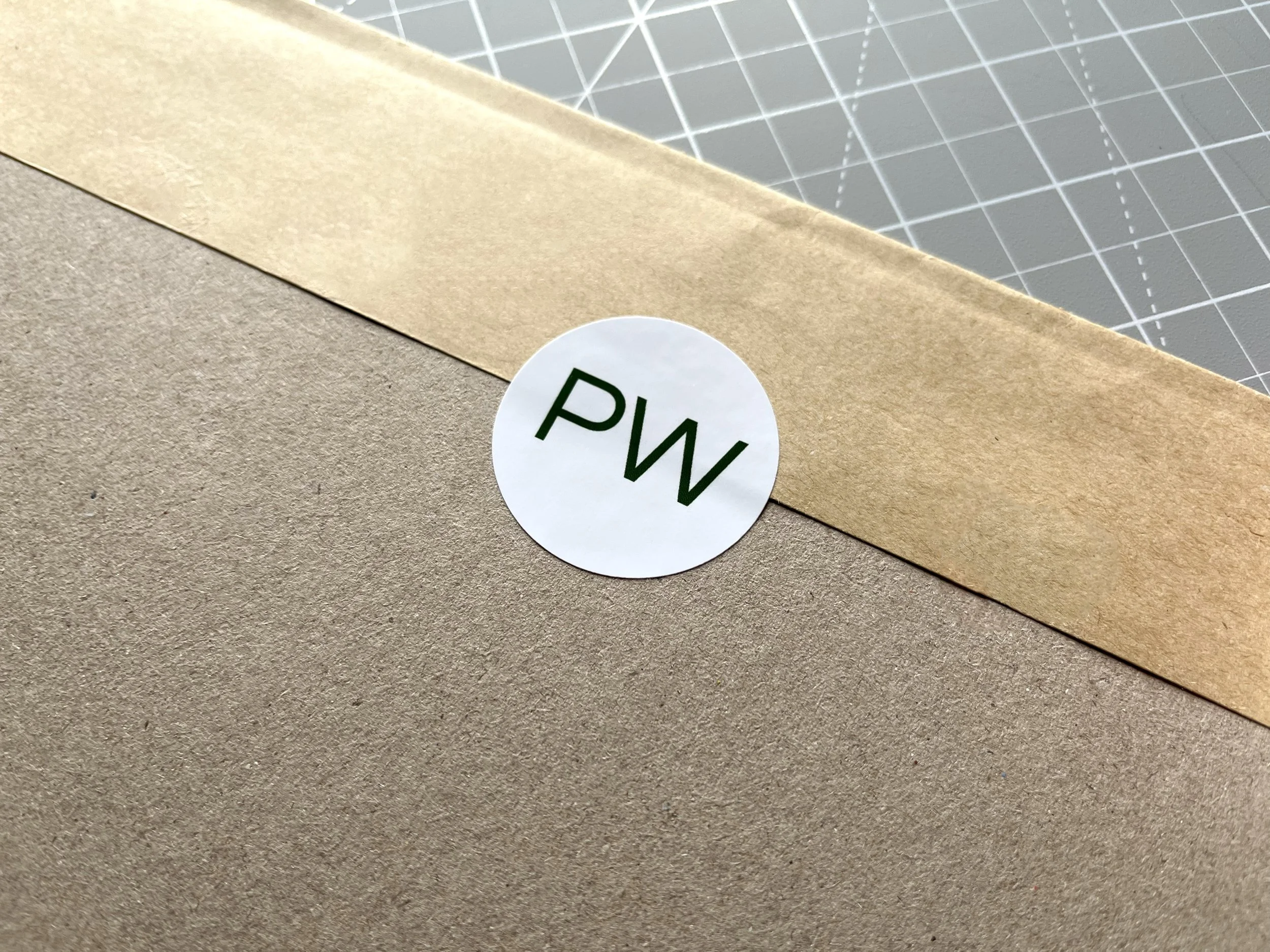 Close-up of a cardboard box sealed with brown packing tape, with a round white label marked 'PW' in green letters, on a cutting mat.