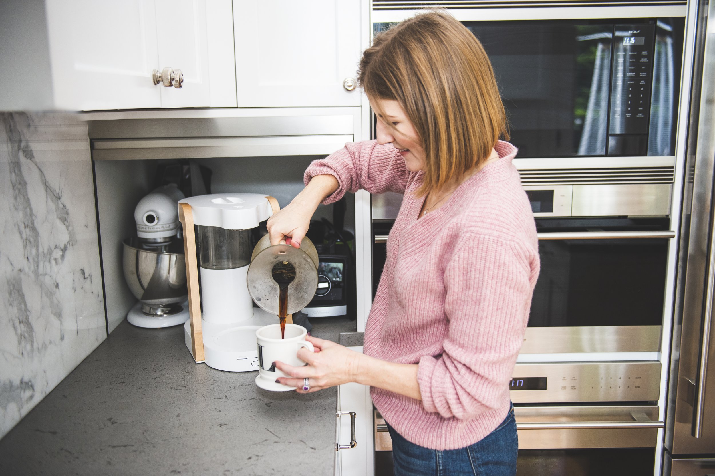 a woman with short dark blonde hair wearing a pink sweater pours coffee into a mug from a Ratio coffee maker