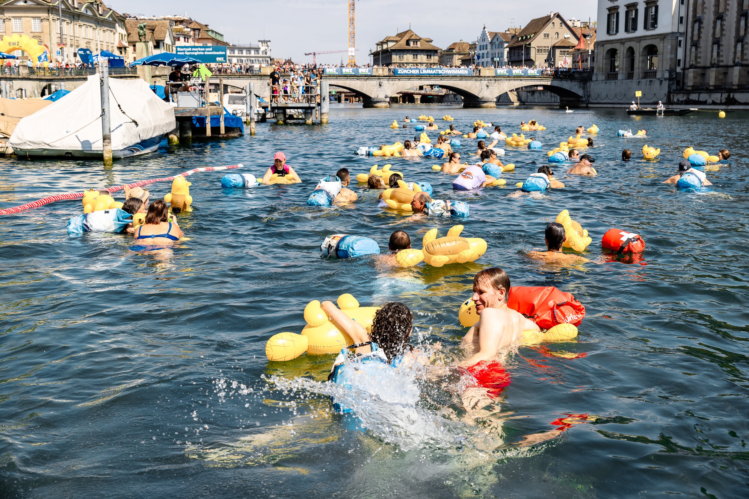 Limmatschwimmen Zürich: swimmers with yellow rubber ducks and dry bags on the Limmat, old town and bridge behind / Schwimmende mit gelben Gummienten und Wickelfischen in der Limmat, Altstadt und Brücke im Hintergrund