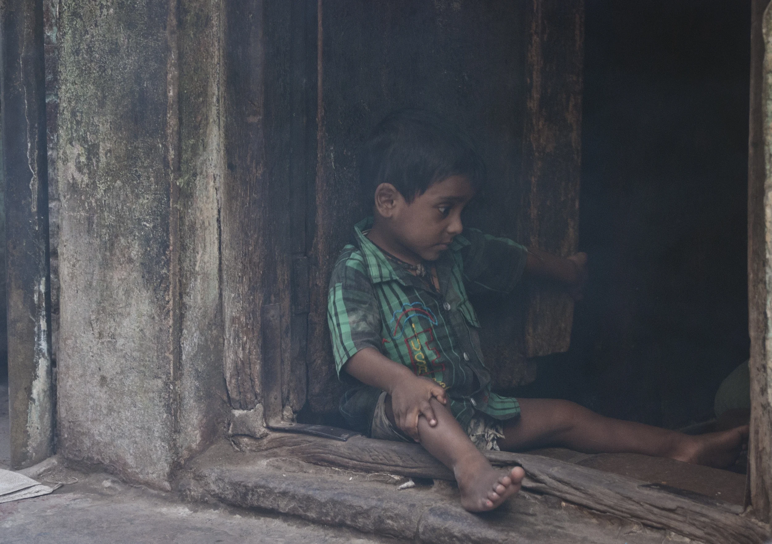 A child acting coy as he sits in the dust filed door way to his home. 