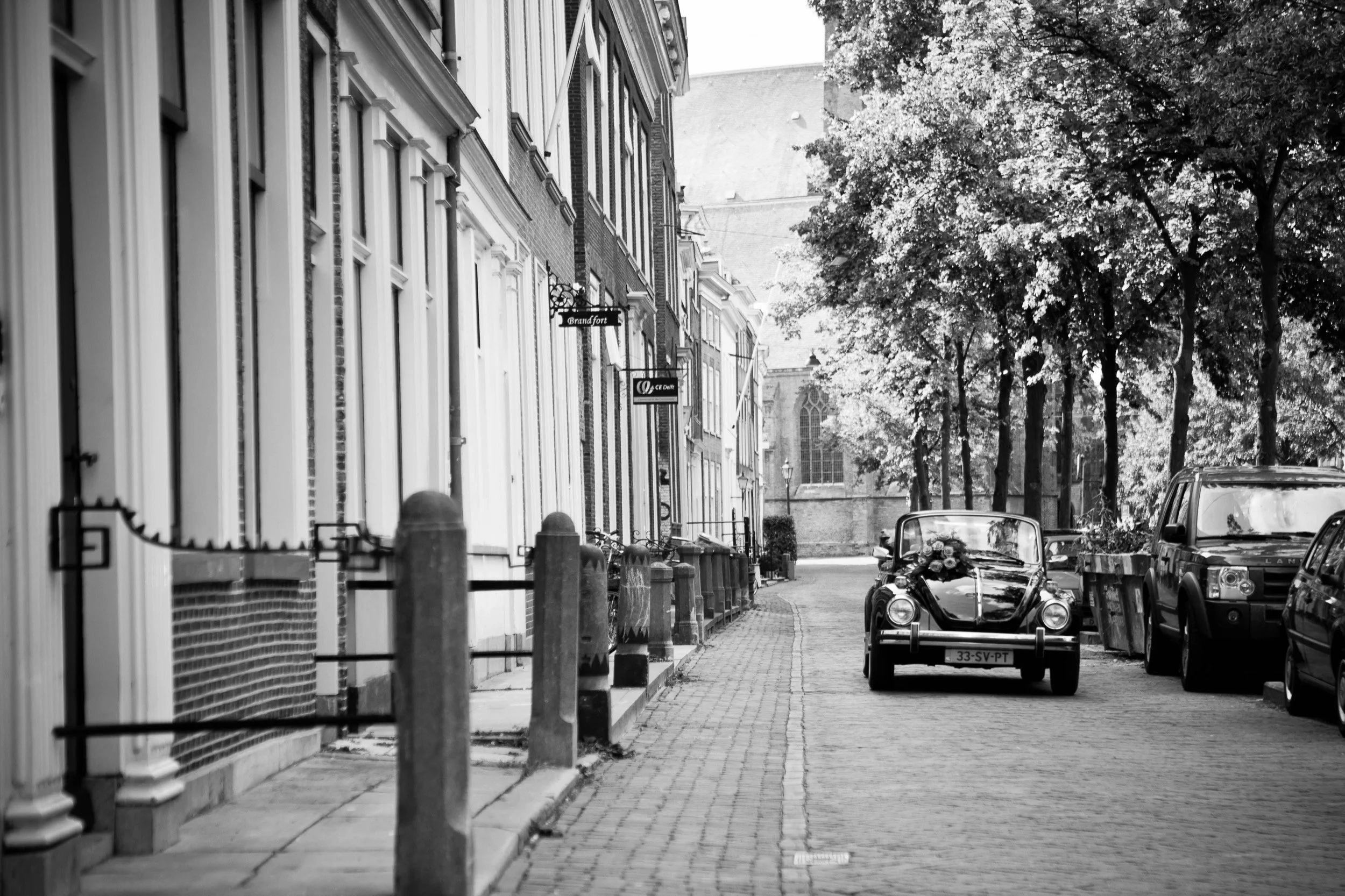 Schets van een straat in een stad met een smalle straat, geparkeerde auto's en een klassieke auto met bloemen, omgeven door bomen en historische gebouwen.
