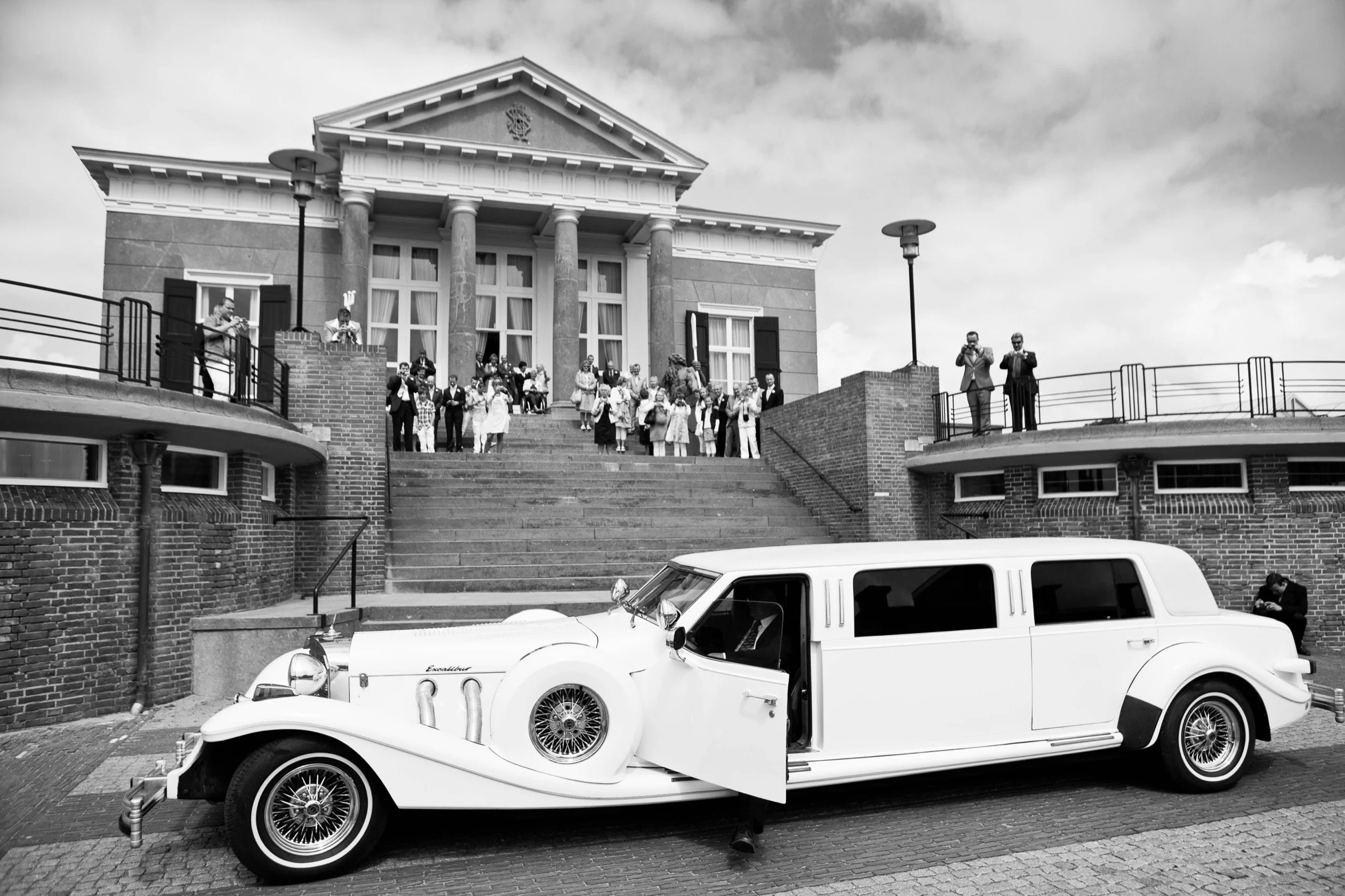 Zwart-wit foto van een klassieke limousine geparkeerd voor een grote witte gebouw met veel mensen op de trap en op de balkonren, die waarschijnlijk deelnemen aan een bruiloft of feest.