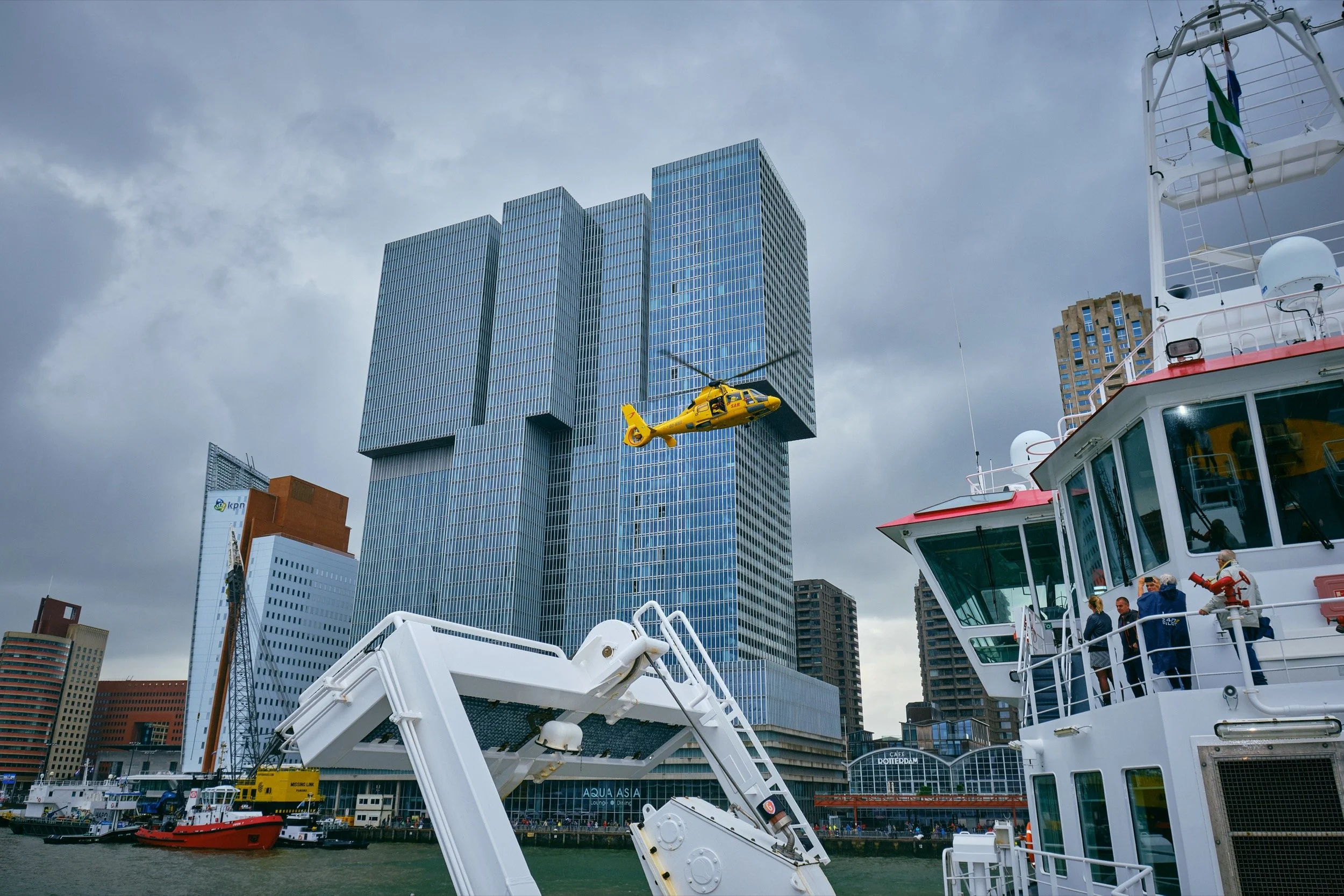 Een gele helikopter vliegt tussen hoge kantoorgebouwen in de stad, gezien vanaf een boot langs de rivier.