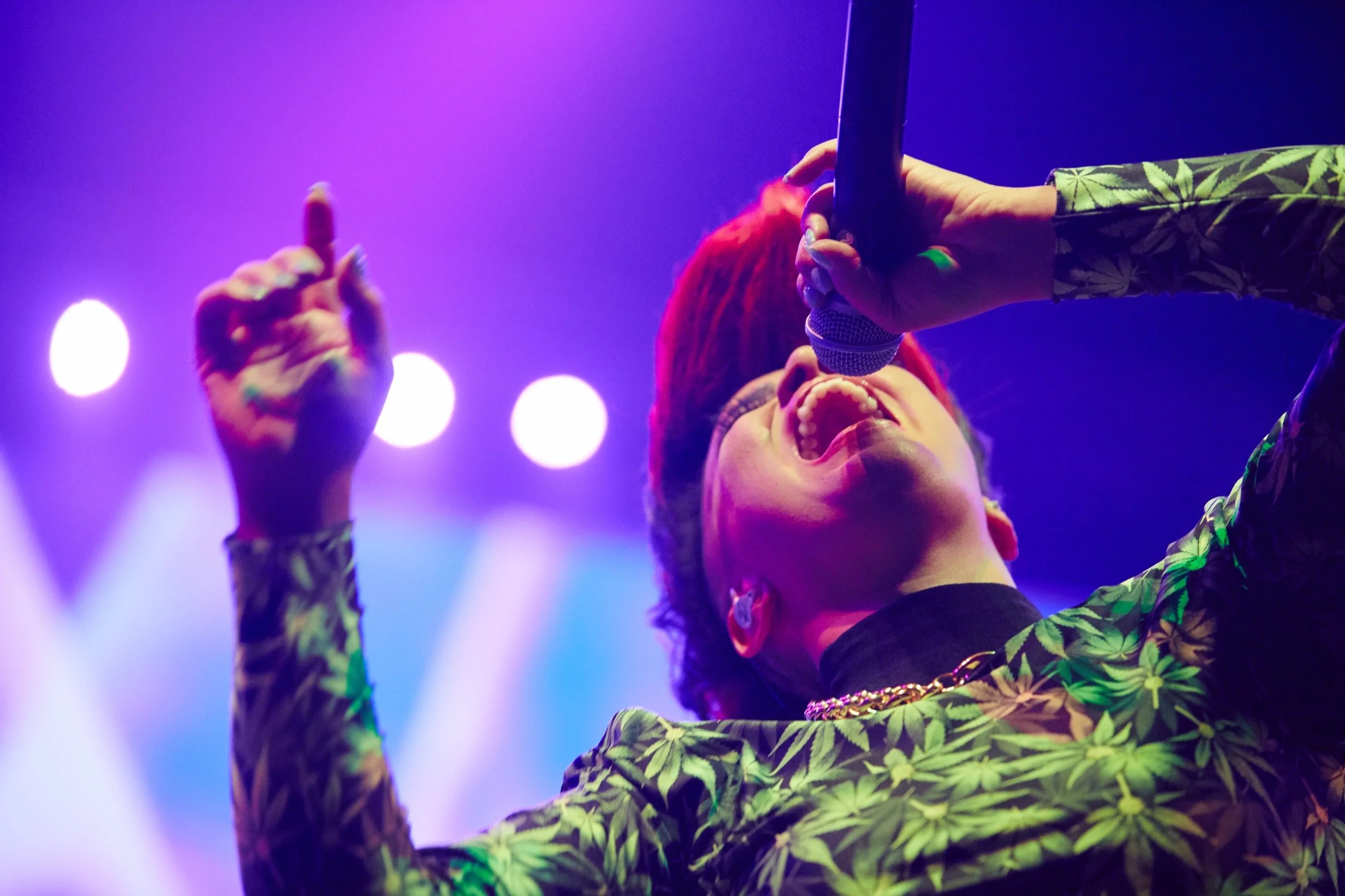 Een zangeres met rood haar en een groene en zwarte bloemenprint shirt zingt met een microfoon onder felle, gekleurde podiumverlichting.