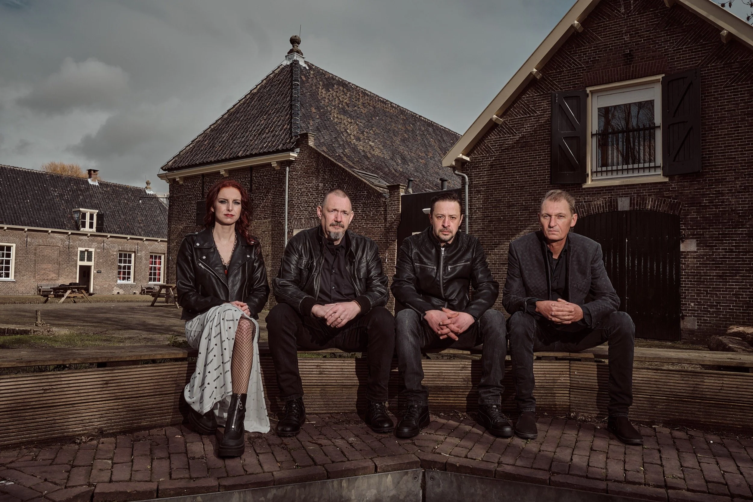 Vrouw en drie mannen zitten op een bank buiten, in een omgevingslandschap met oude bakstenen gebouwen. Ze dragen donkere kleding en kijken serieus.