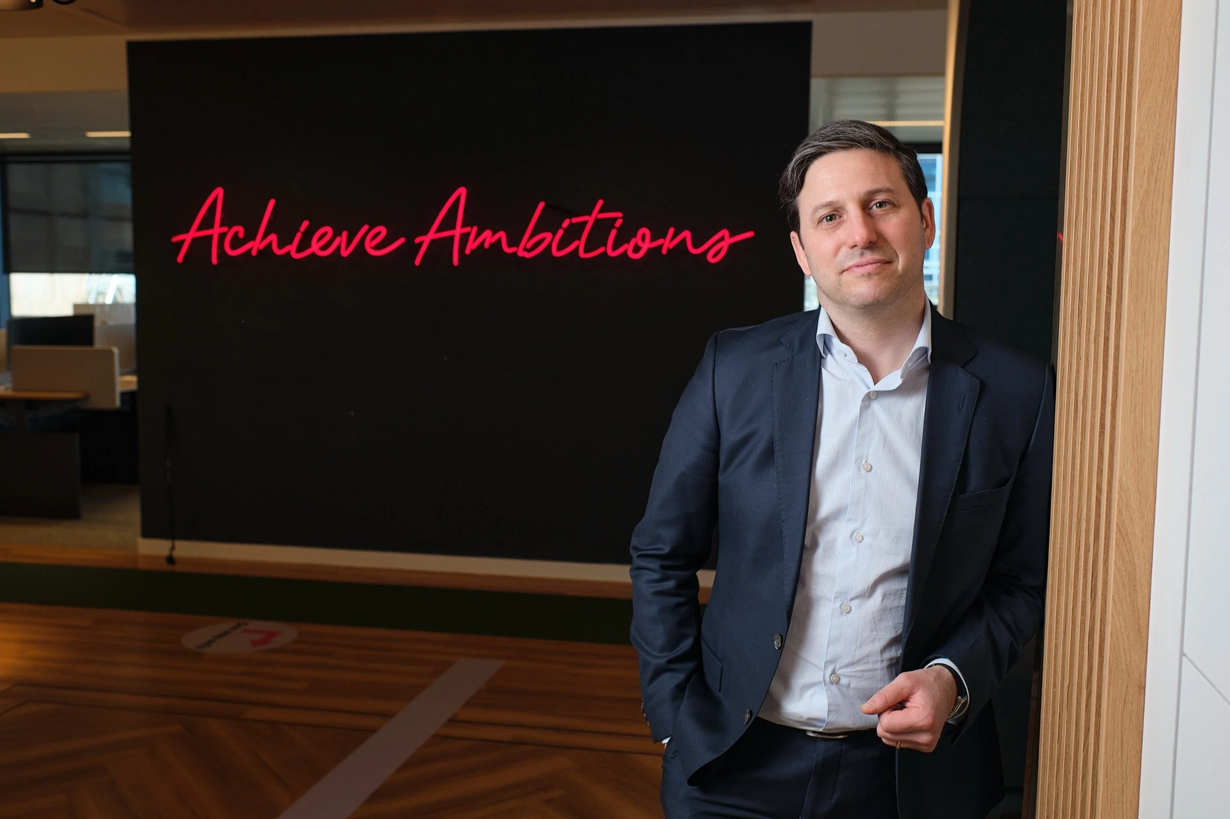 Man in een blauw pak staand bij een houten muur, met een blackboard op de achtergrond met de woorden 'Achieve Ambitions' in roze Schrift.
